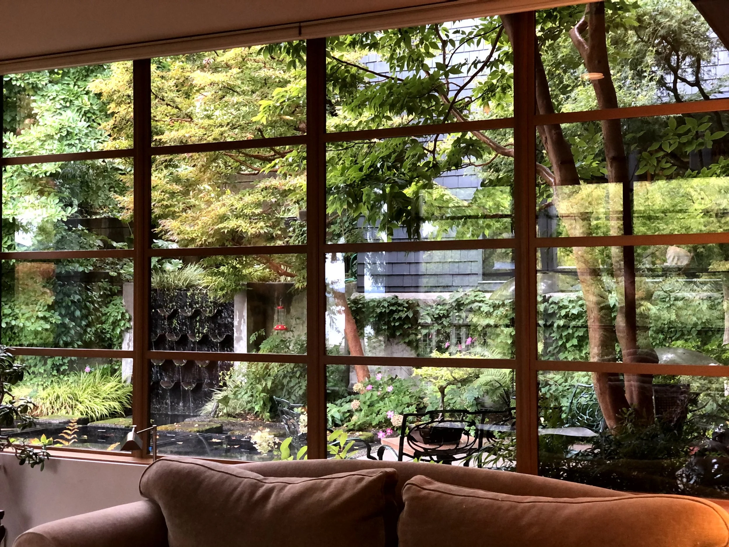 View of a lush green garden through large window panes, with trees, plants, a fountain, and outdoor furniture visible outside; inside has a sofa.