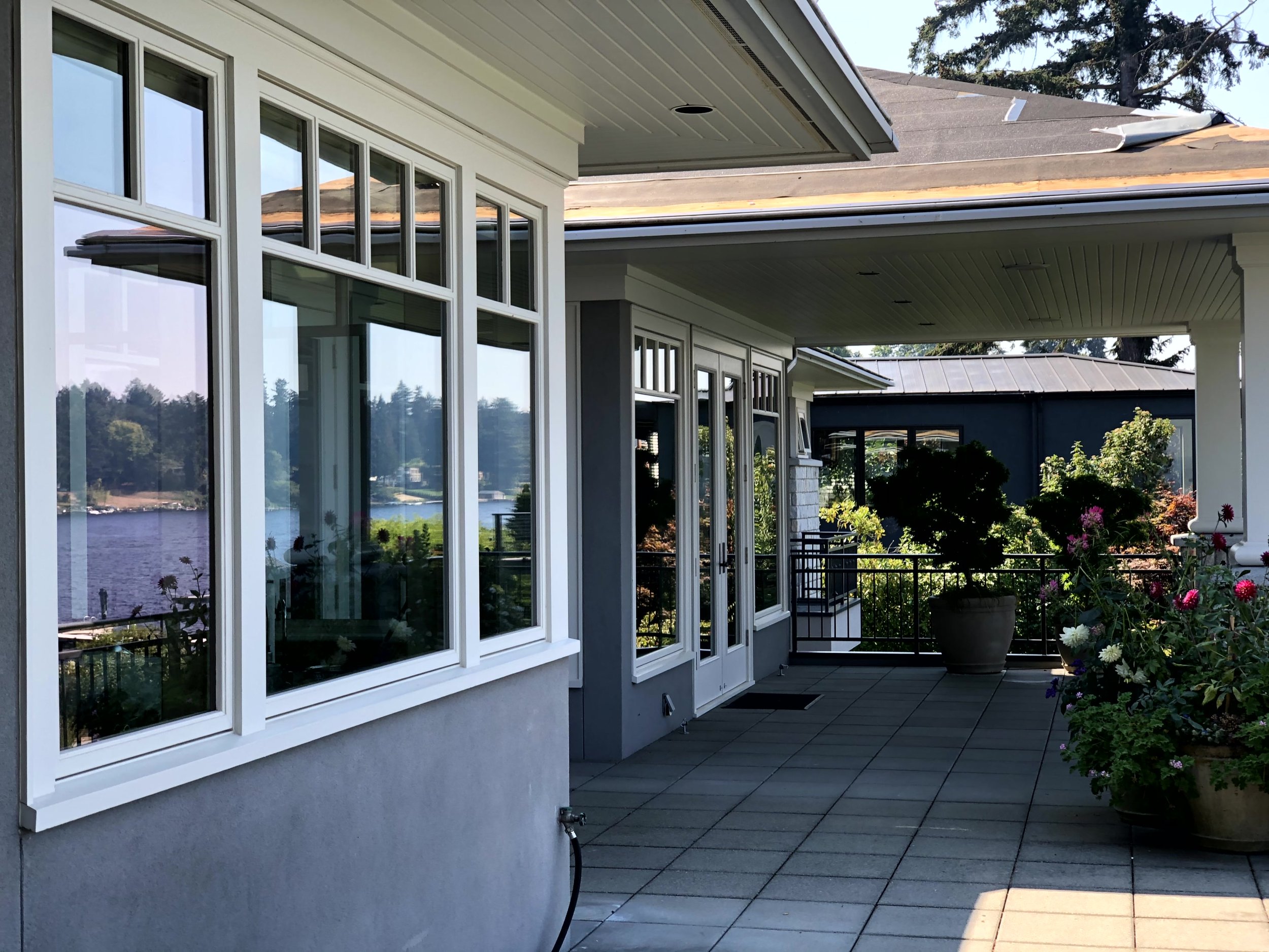 A covered outdoor patio area beside a house with large windows, potted plants, and a view of a river in the background.