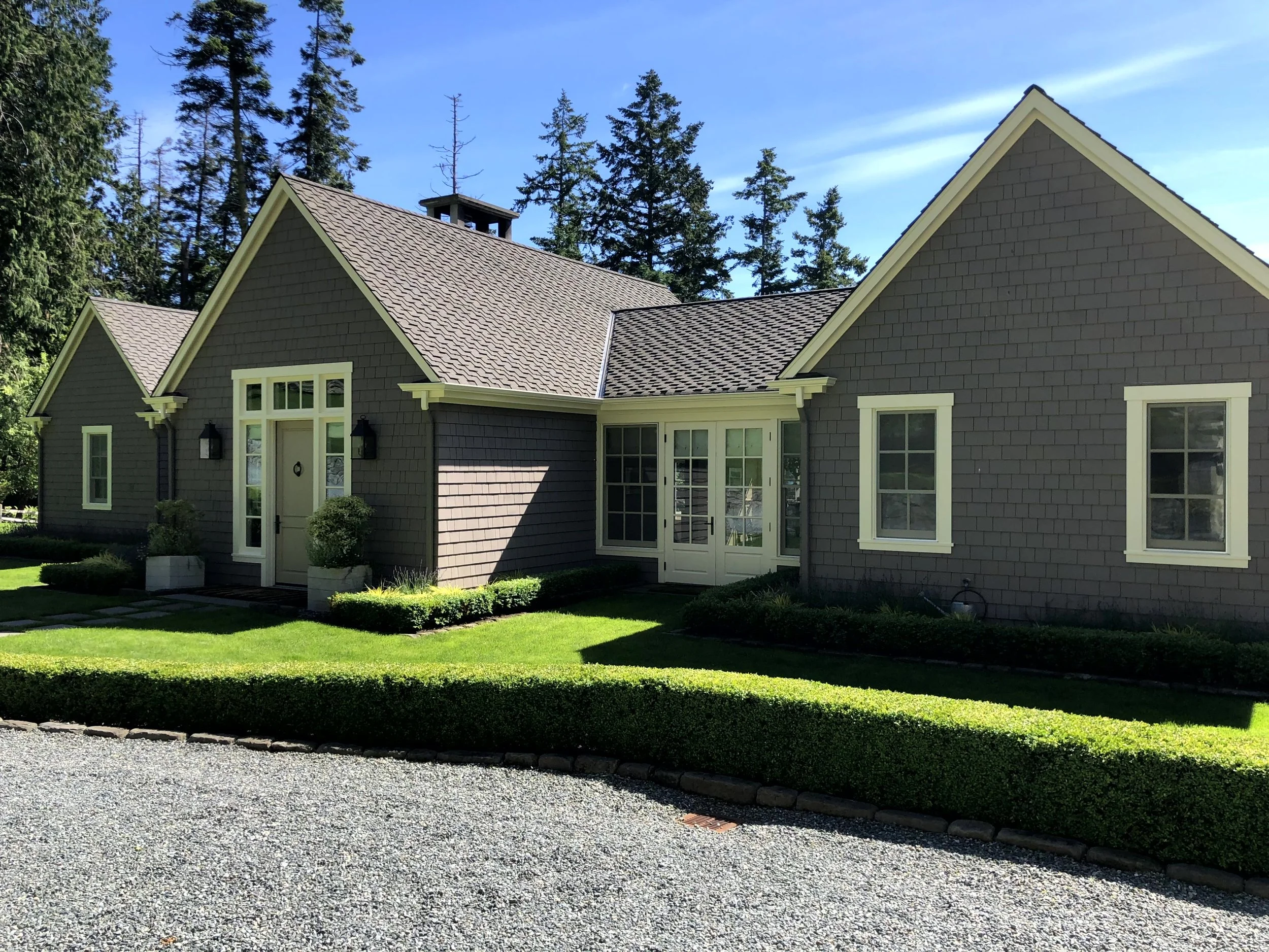 A modern house with gray exterior, gabled roof, and white trim, surrounded by green lawn and bushes, with a gravel driveway in front, and tall trees in the background under a clear blue sky.