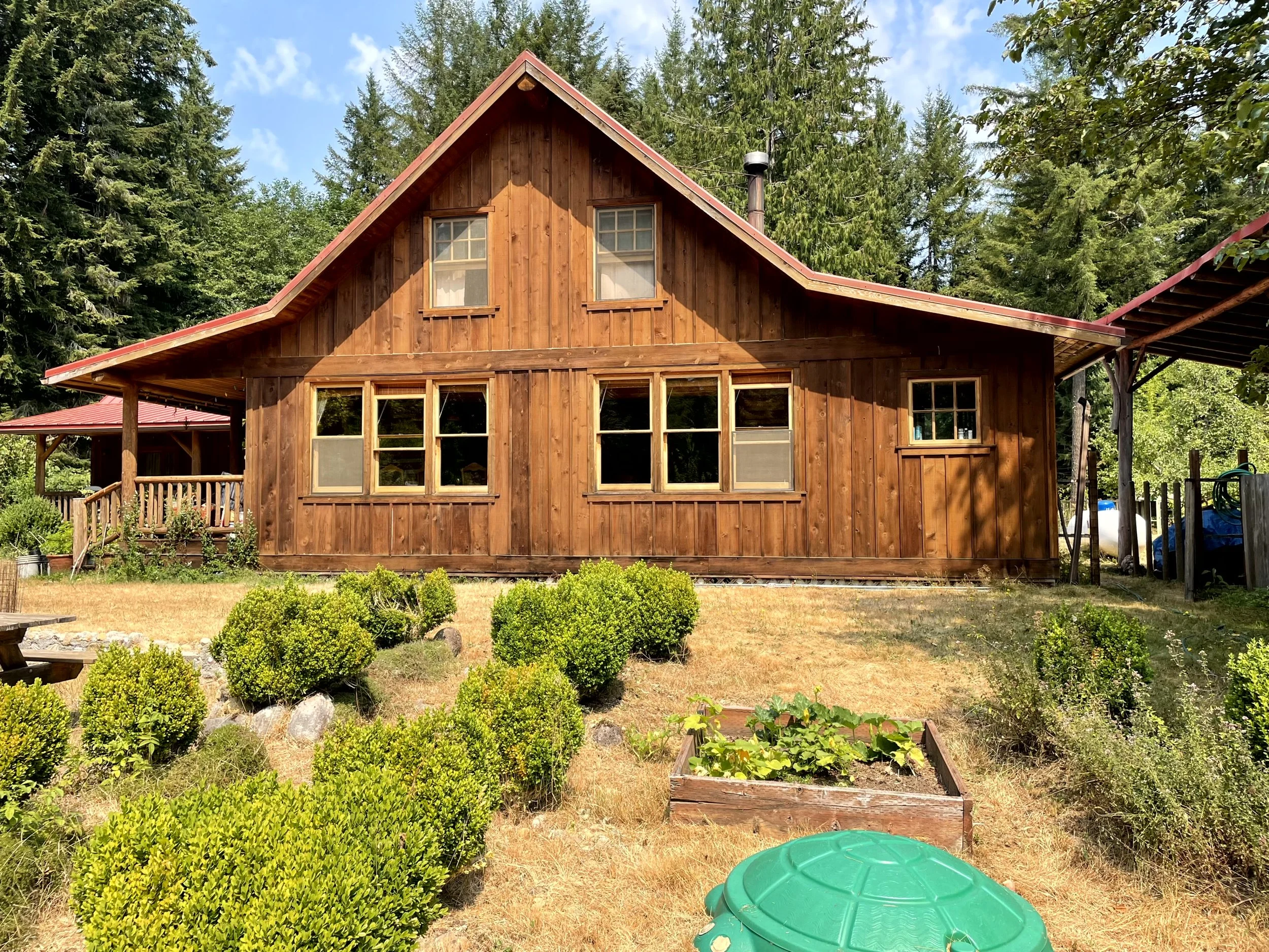 A wooden house with a gabled roof and multiple windows, surrounded by a garden with bushes and plants, in a forested area with trees and a partly cloudy sky.