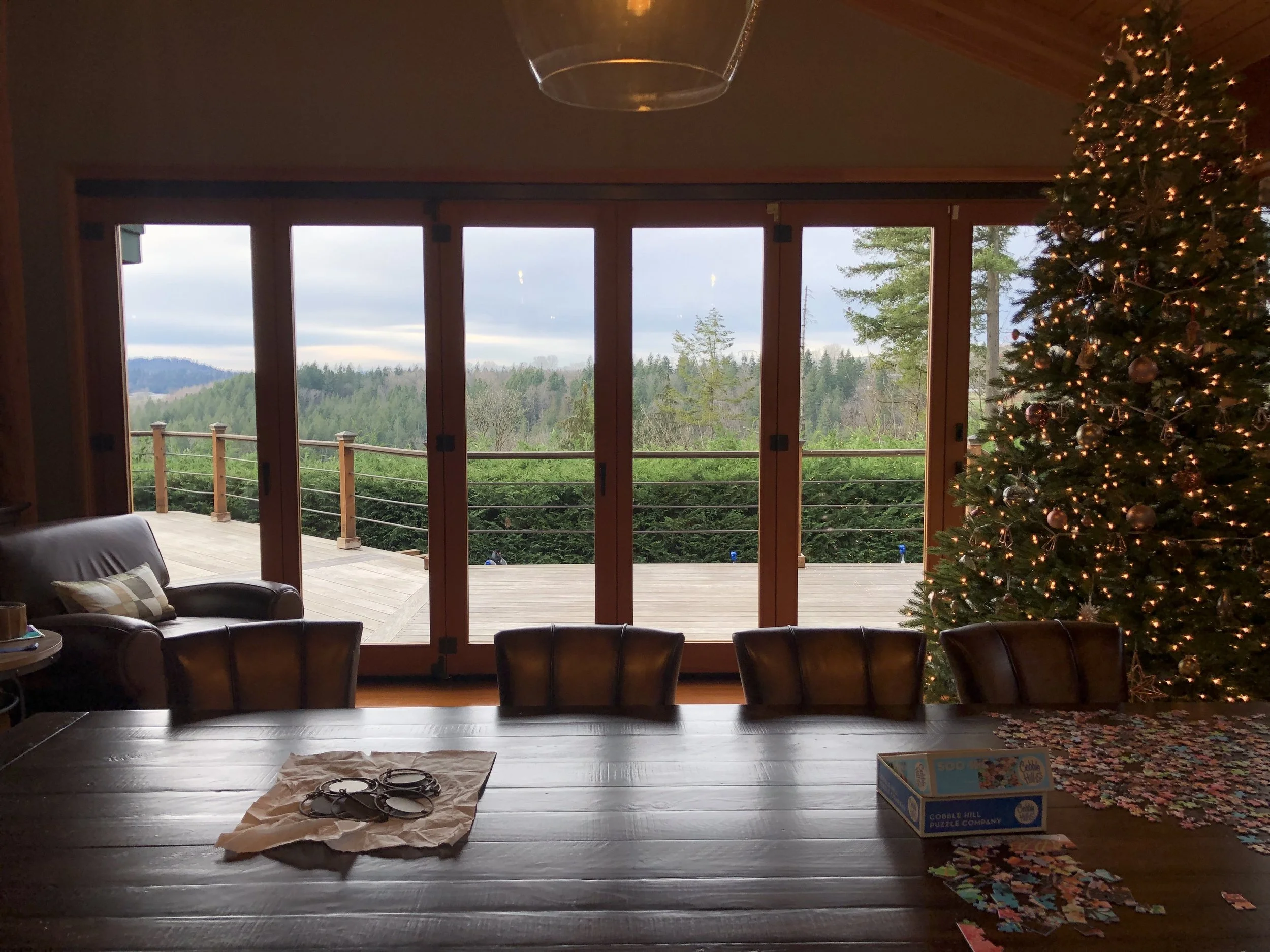 Interior of a dining area with a large wooden table, four chairs, a box of puzzle pieces, a partially completed puzzle, a Christmas tree decorated with lights and ornaments, and a view of a deck and forest outside through large sliding glass doors.