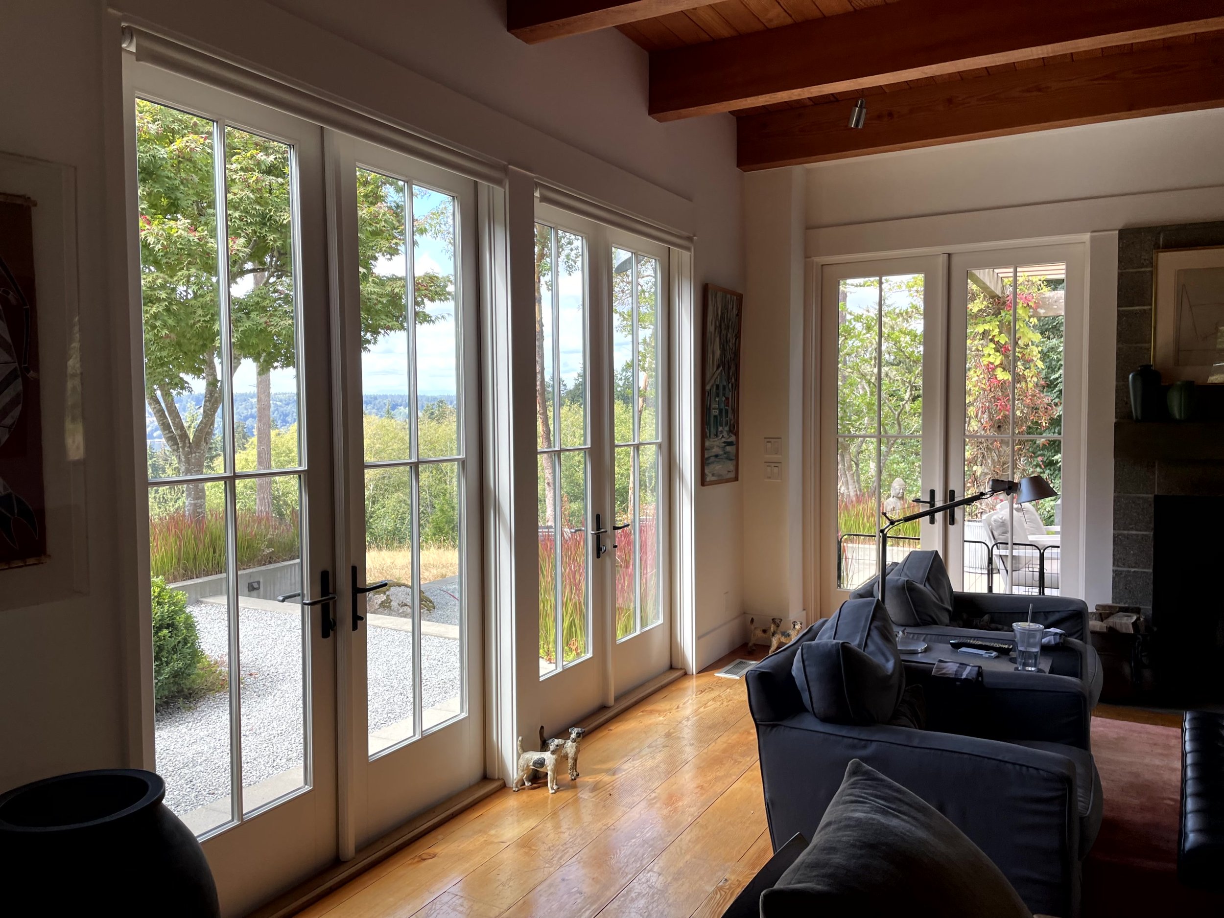 Living room with large windows showing greenery outside, dark blue sofas with cushions, a lamp, and a small dog figurine near the door.