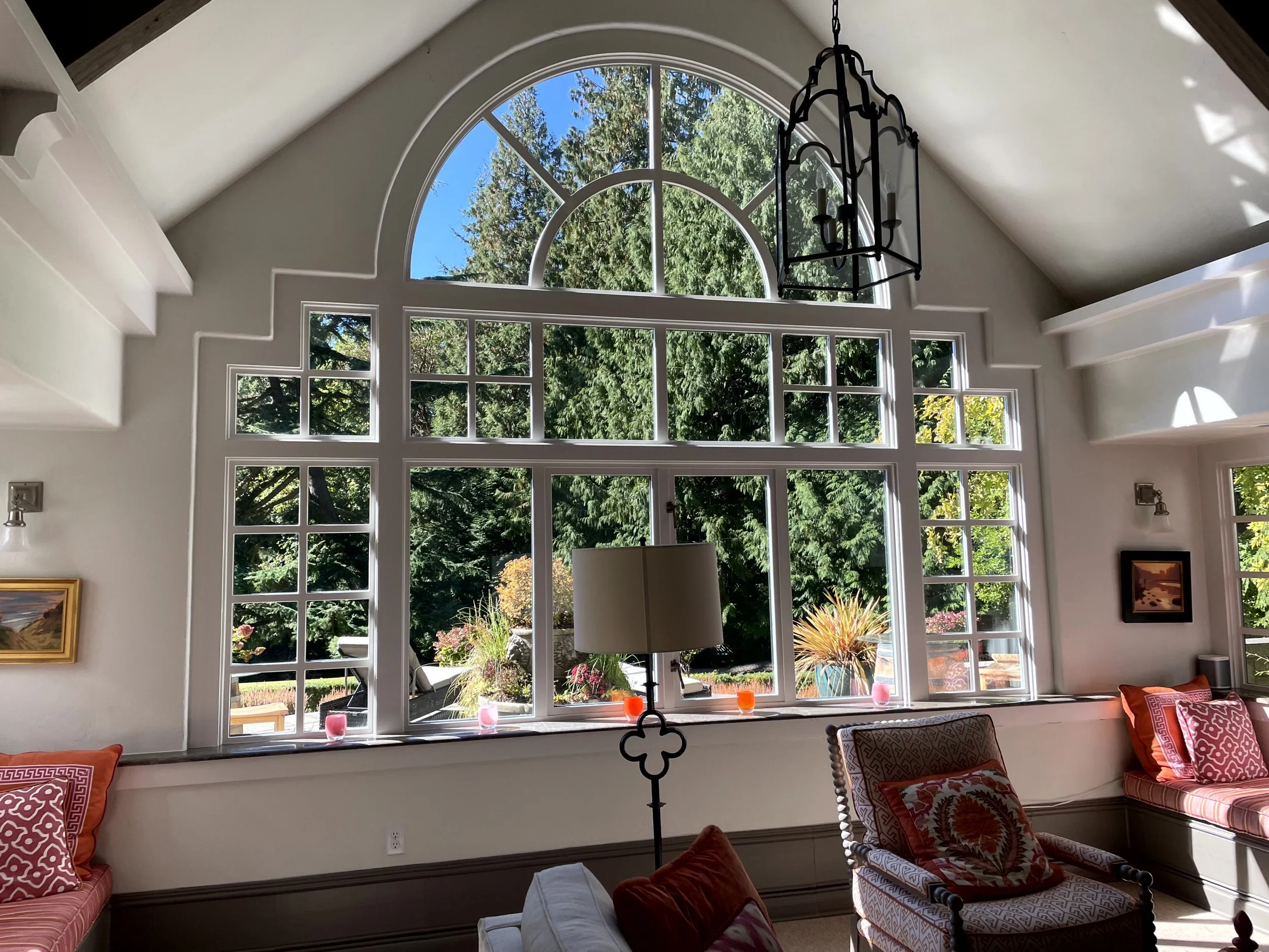 Living room with large arched window revealing greenery outside. Decorative pillows on seating, chandelier hanging from ceiling, and plants on windowsill.