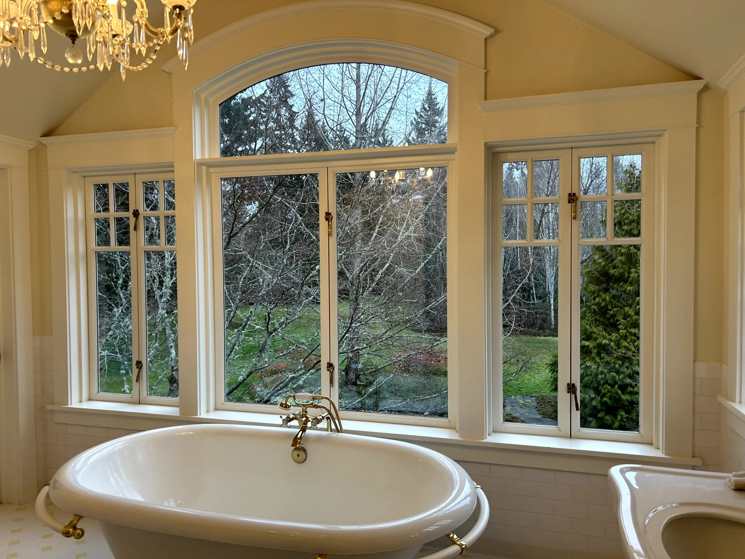 Oval bathtub with brass fixtures in front of large multi-paneled window showing a winter landscape with leafless trees and green grass.
