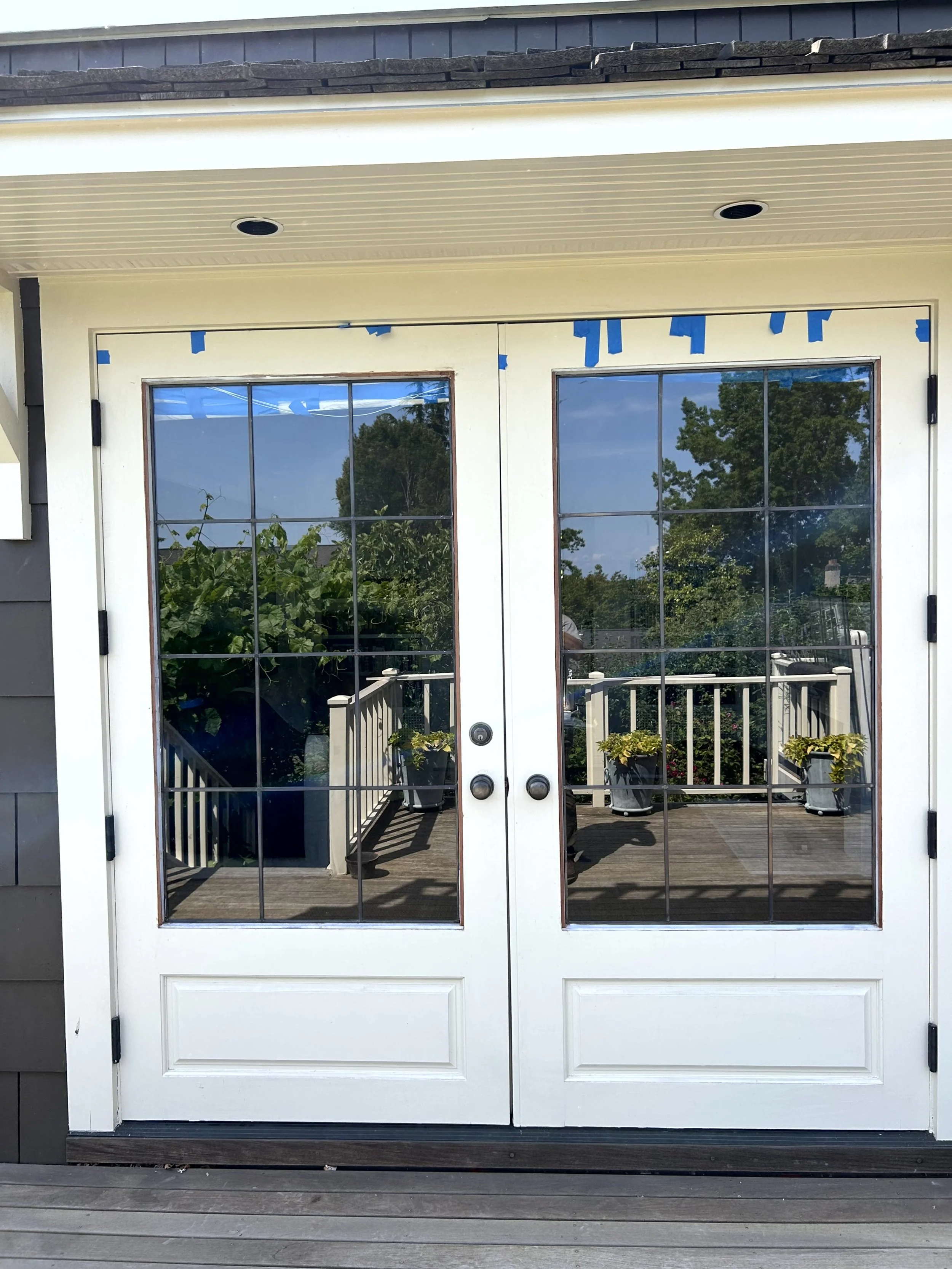 Double glass doors with a wooden deck outside, potted plants on the porch, and trees reflected in the glass.