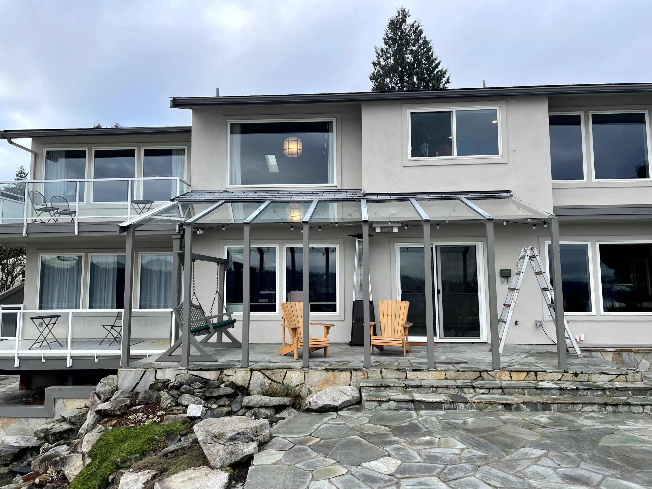 Back view of a modern multi-story house with large windows, a stone patio, and a glass-covered porch with outdoor furniture, including two Adirondack chairs, a swing, and a ladder nearby.