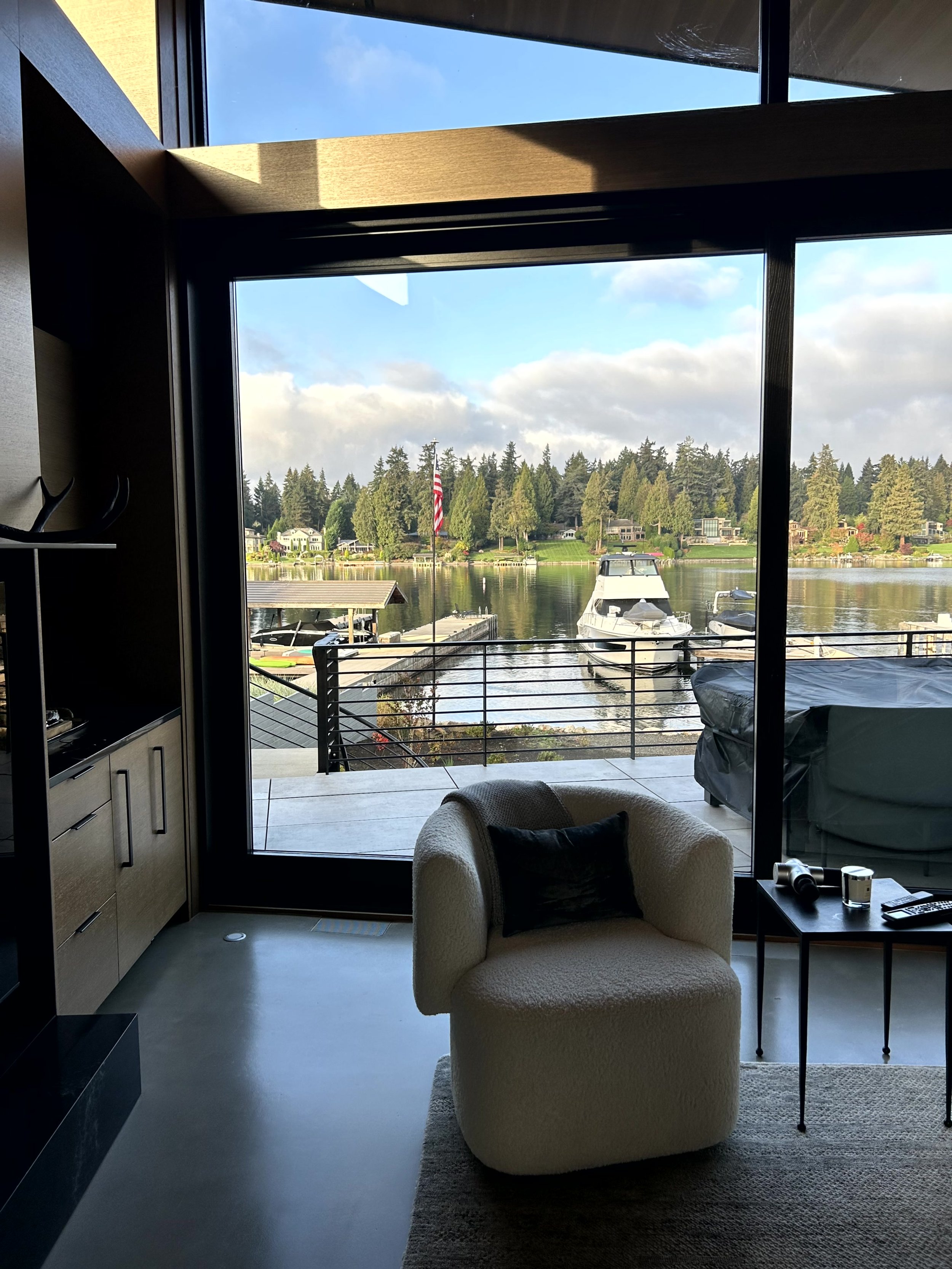 Living room with a large glass window overlooking a marina with boats, trees, and an American flag in the background.