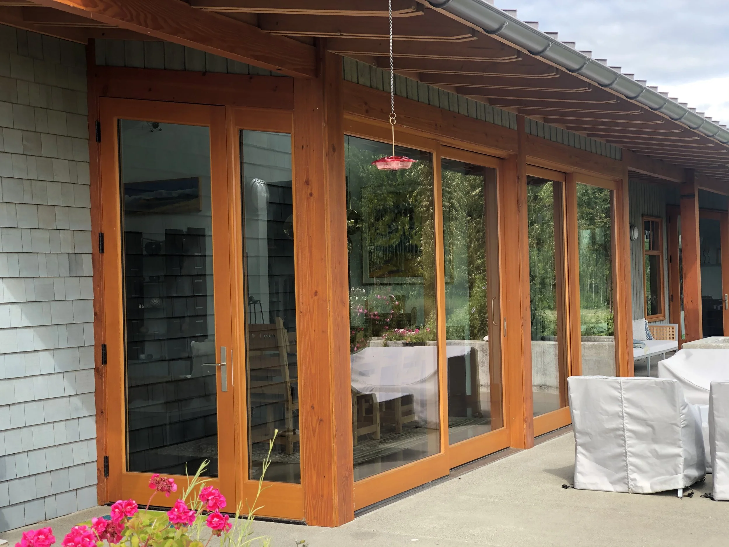 Exterior view of a house with large glass sliding doors and wooden framing, outdoor patio with white covered chairs, pink flowers in the foreground, and a lush green garden visible through the glass.