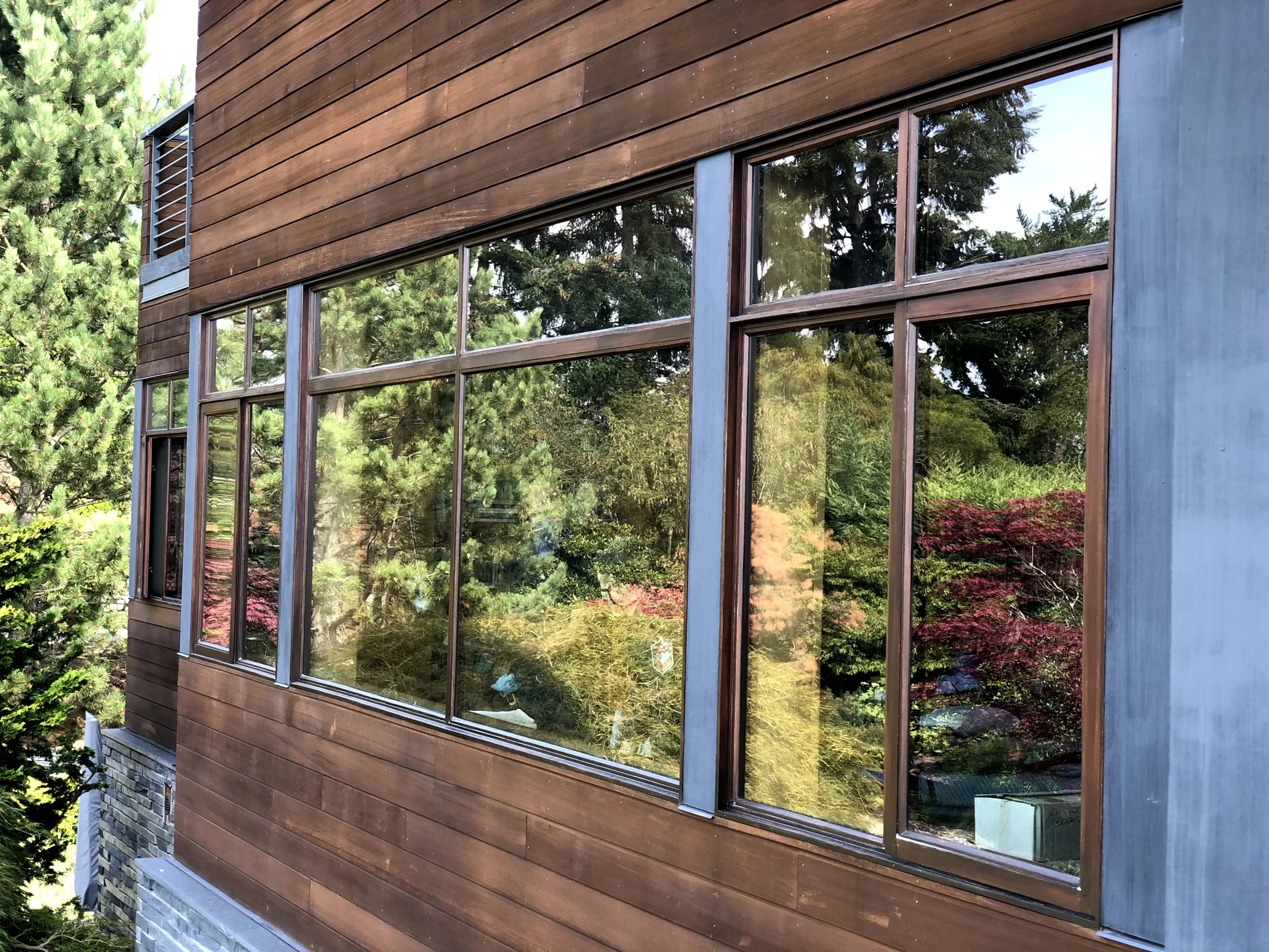 The image shows a modern house with large glass windows, wooden siding, and surrounding trees and greenery.