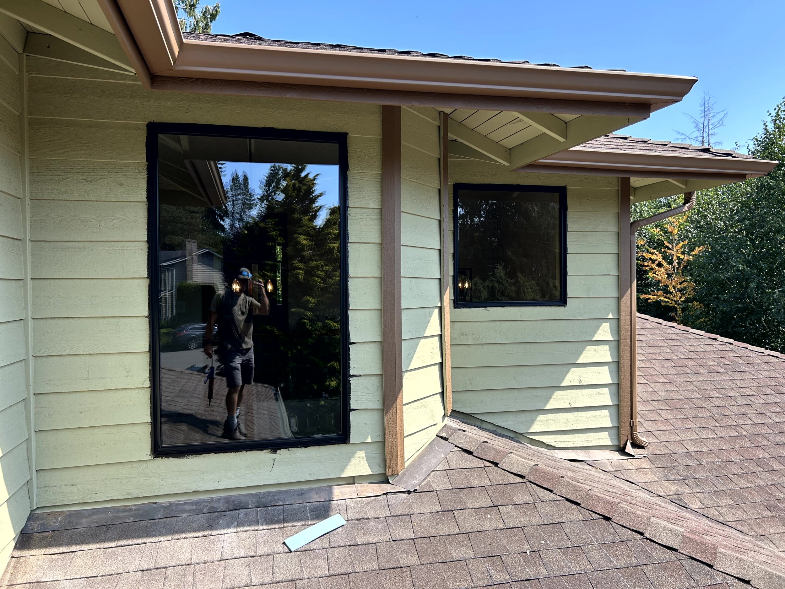 View of a house’s exterior showing two large rectangular windows on a yellowish-green wooden exterior wall, with reflections of trees and a person taking a photo, and a sloped shingled roof under a clear blue sky.