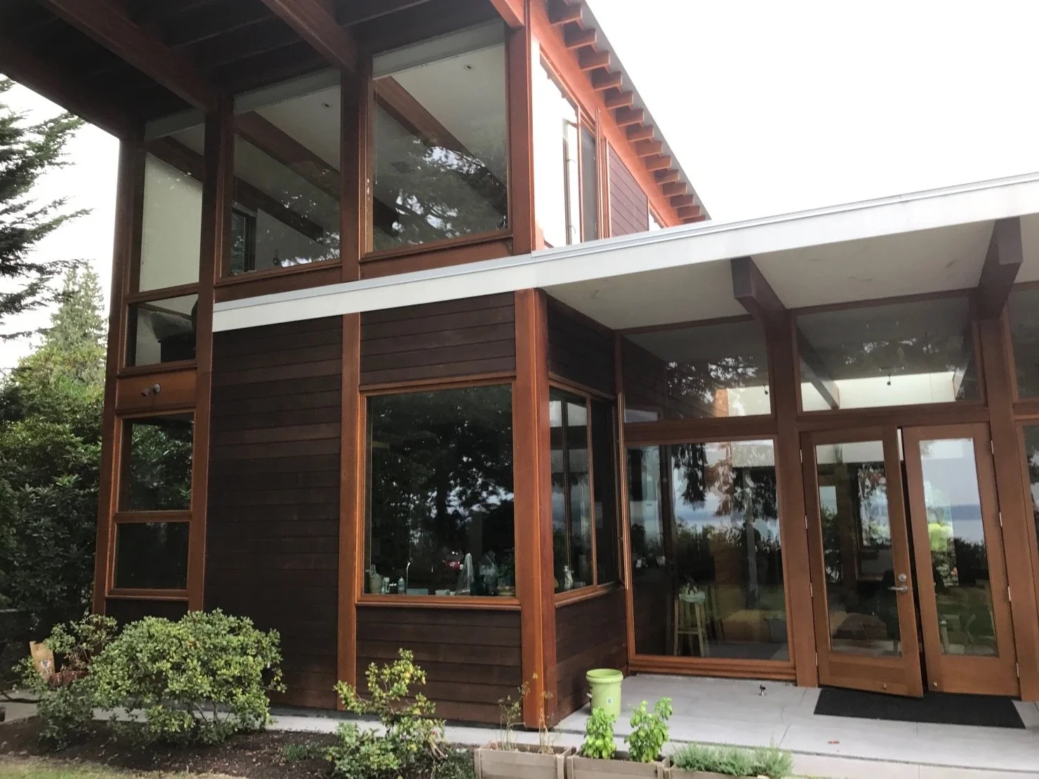 A two-story house with large glass windows and wooden siding, surrounded by greenery and trees.