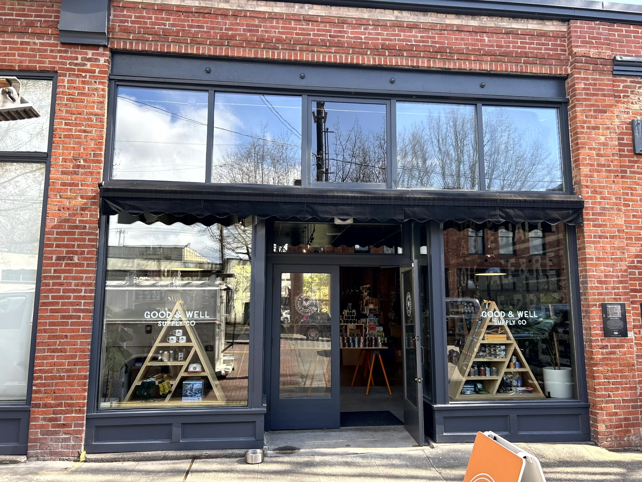 The storefront of 'Good & Well Supply Co.', a retail shop with large glass windows and a glass door, displaying shelves with products inside. The shop is in a brick building with a black awning above the entrance.