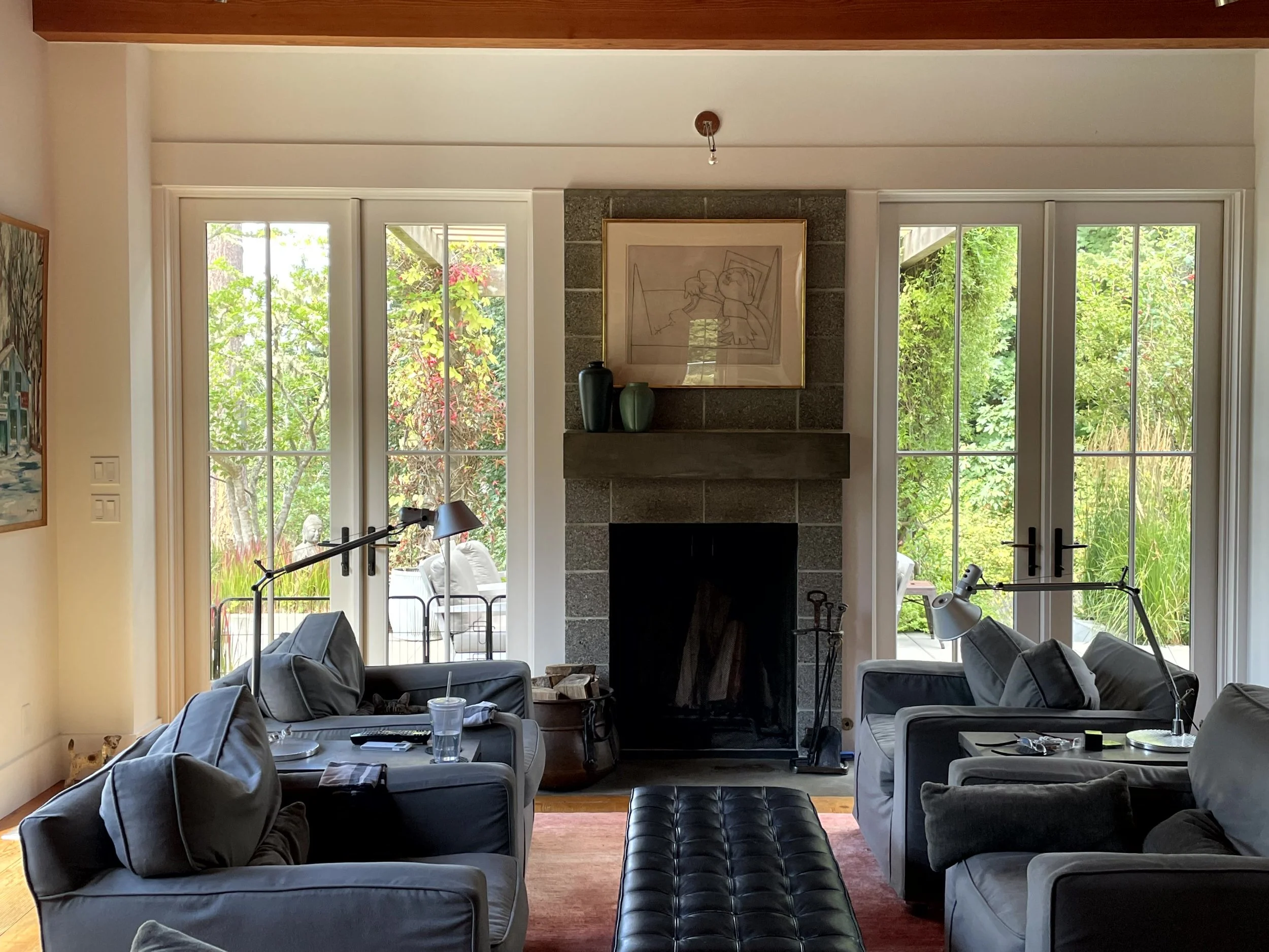 Living room with gray sofas, a black leather ottoman, a fireplace with artwork above, and large windows showing a lush outdoor garden.