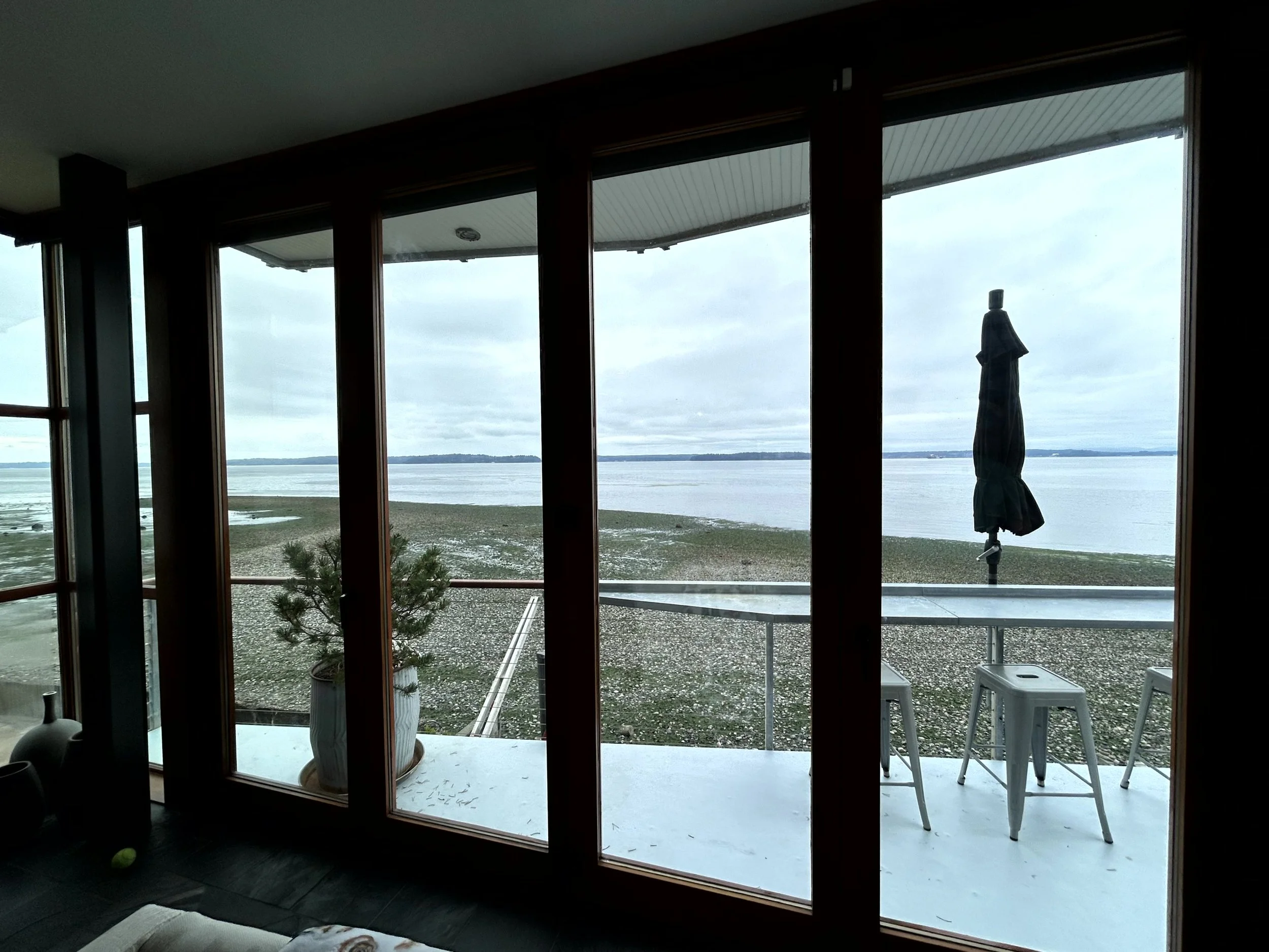 View through large glass doors showing a beach with pebbles and a calm body of water under a cloudy sky, with a closed outdoor umbrella and three white chairs on a patio.