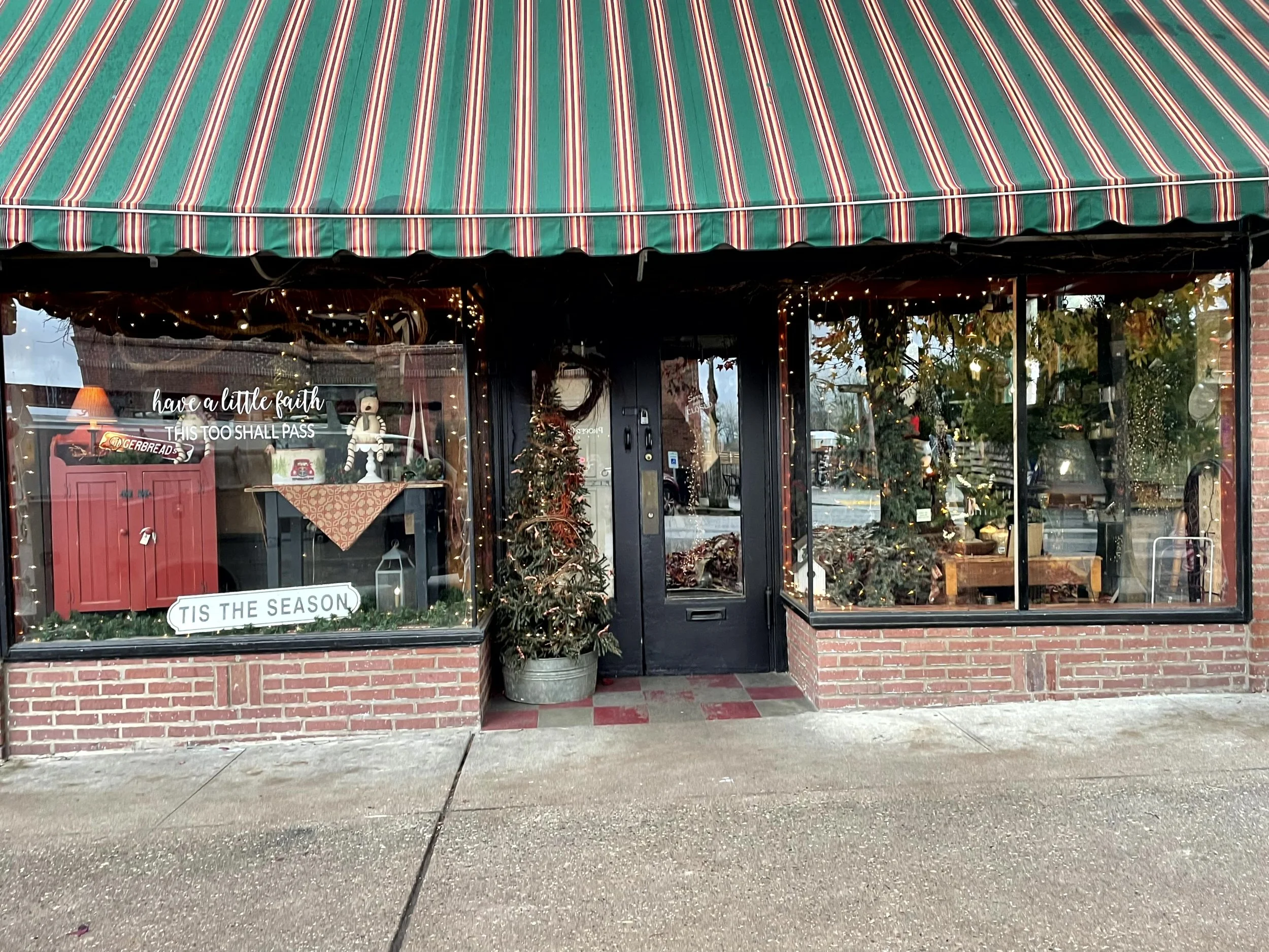 A storefront decorated with Christmas trees and holiday decorations inside, with signs that say 'have a little faith this too shall pass' and 'TIS THE SEASON'. The shop has a striped red, green, and white awning, large glass windows, and a brick exte