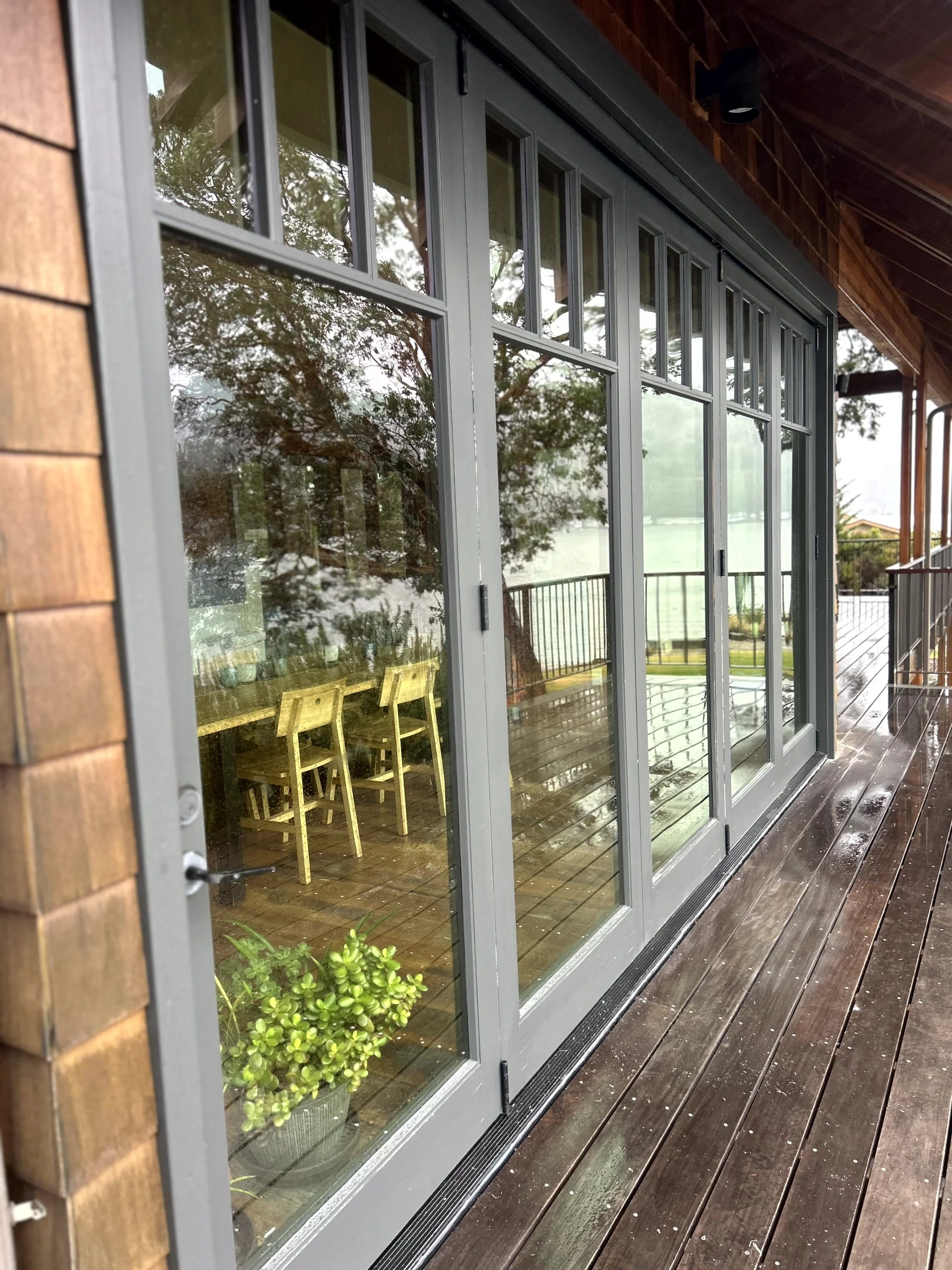 View of a house's large glass sliding doors with reflections of trees outside, showing a wooden deck and interior with a dining table and chairs, and a plant inside.