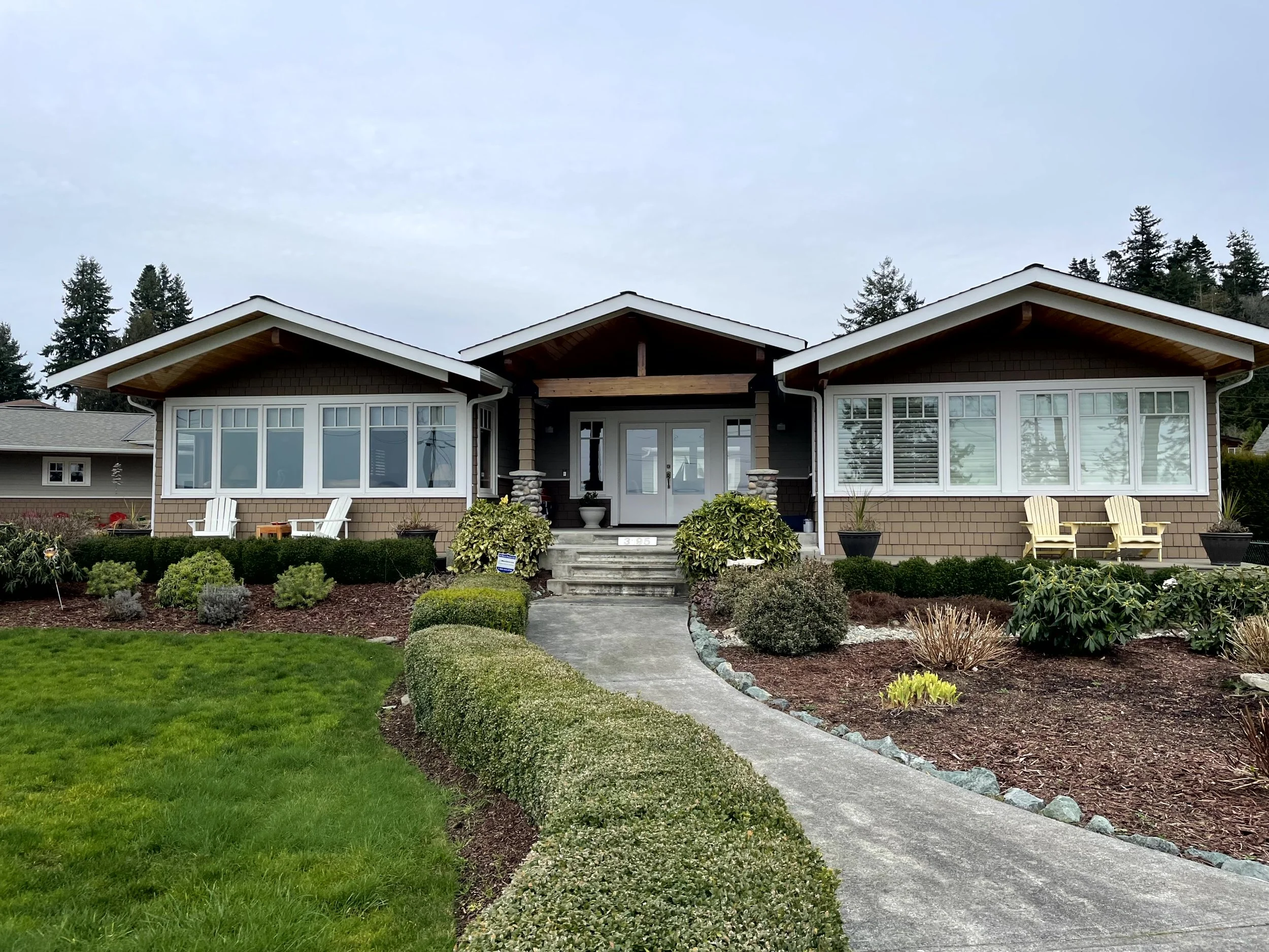 Front view of a modern single-story house with a landscaped yard, concrete walkway, and large bay windows on each side of the entrance, with trees in the background.