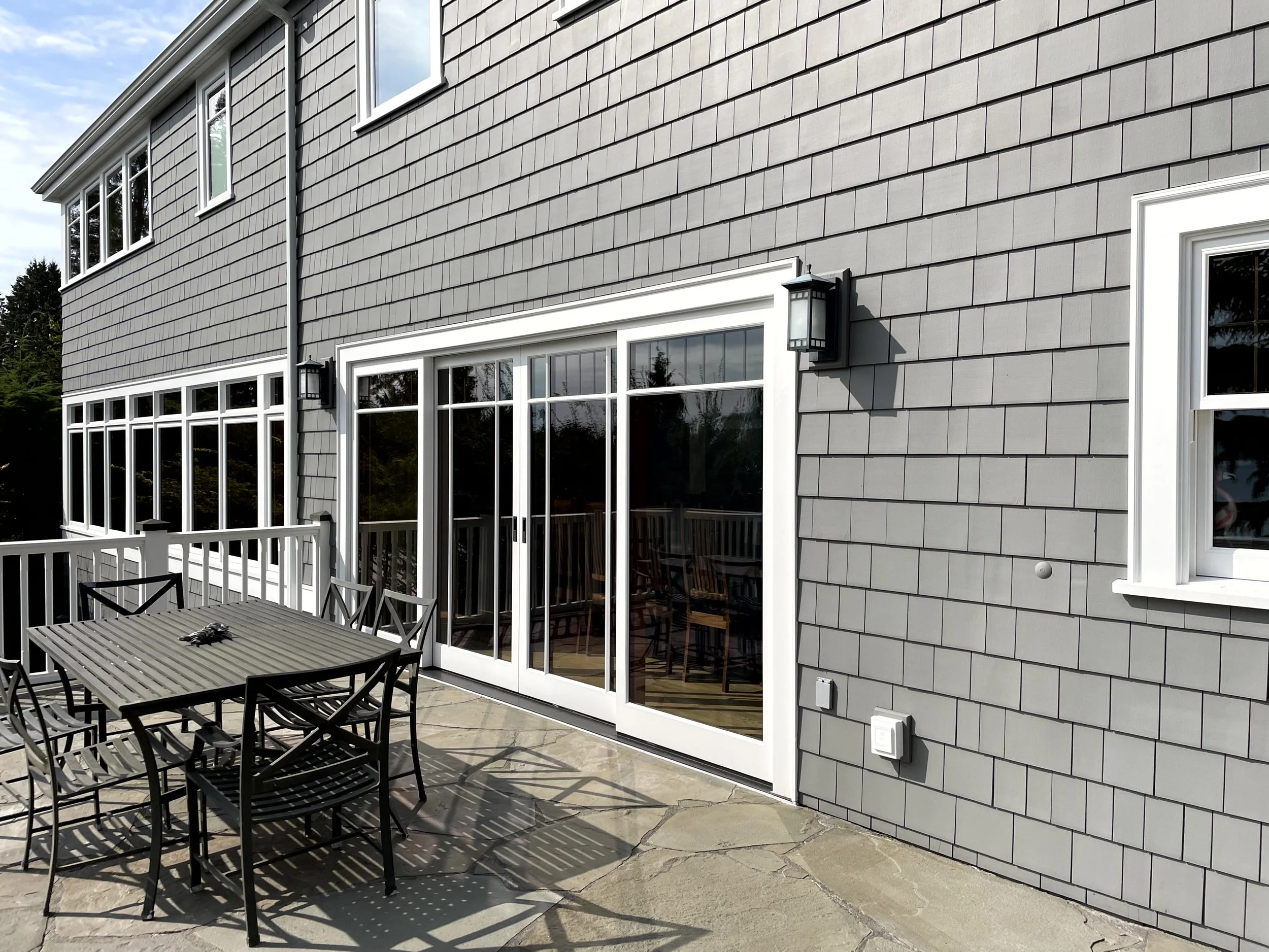 Backyard patio with black metal table and chairs, sliding glass door, gray shingled house exterior, outdoor wall sconces, and a railing with trees in the background.