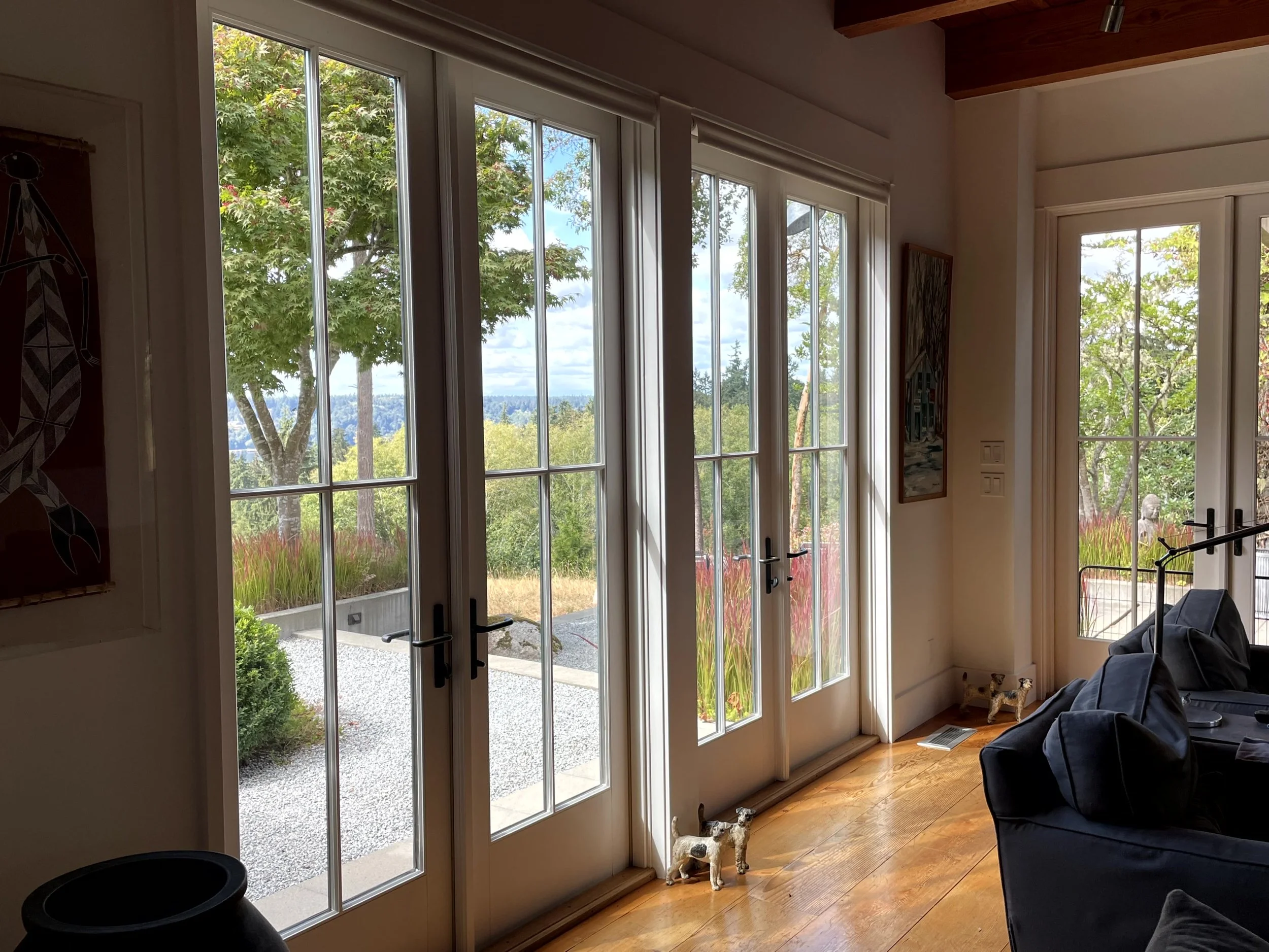 Living room with large windows and glass doors showing outdoor greenery and trees, with hardwood flooring and framed artwork on the wall.