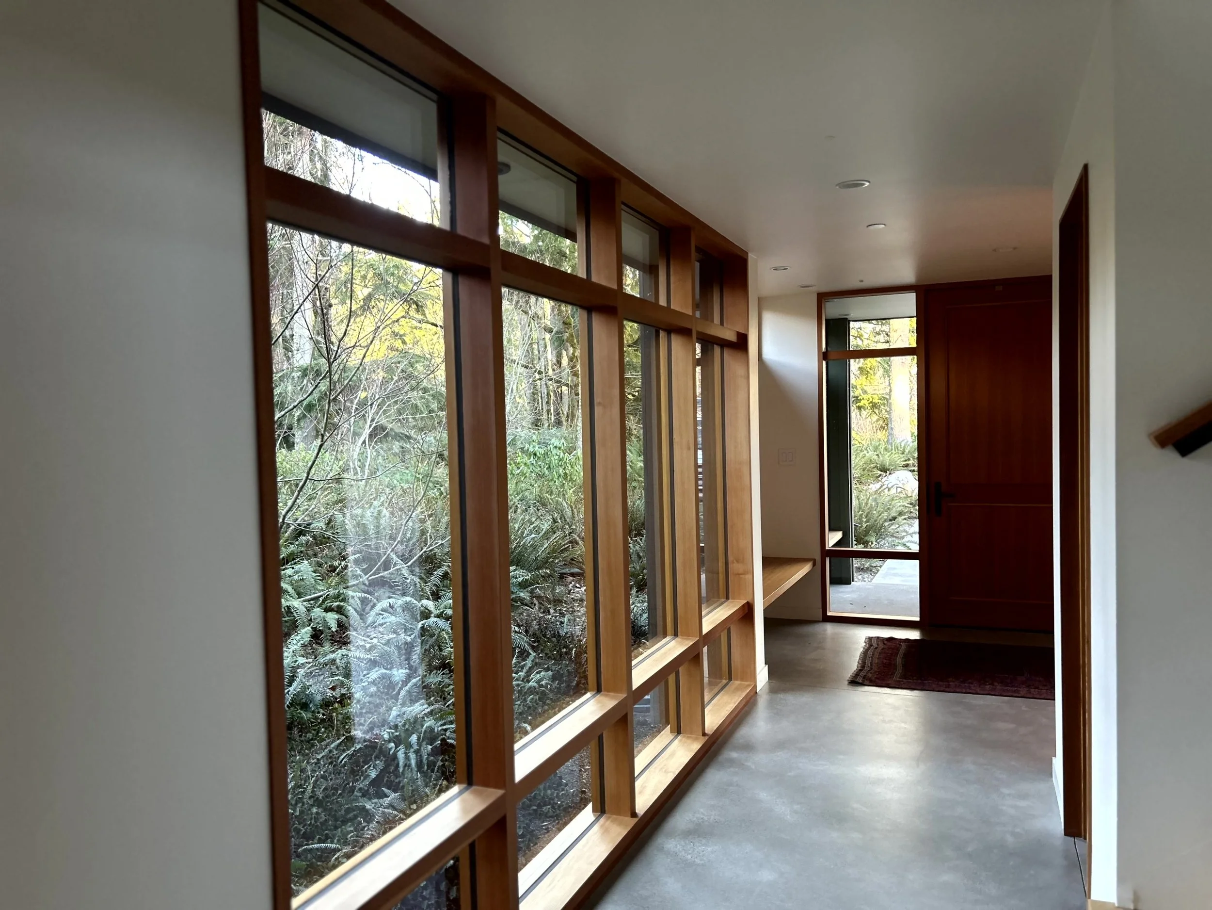 Interior view of a modern house with large glass windows showing a wooded area outside, wooden door, and a small carpet on a concrete floor.