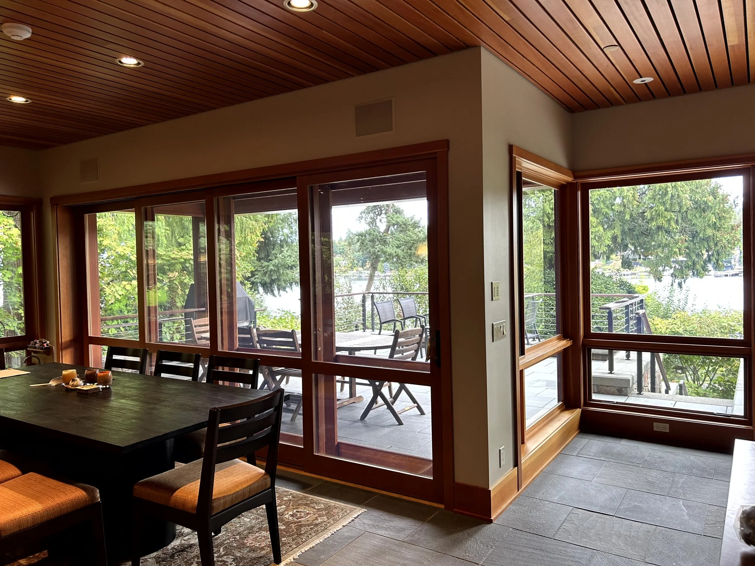 Indoor dining area with large windows overlooking a deck and a lake, wooden ceiling, and tiled floor.