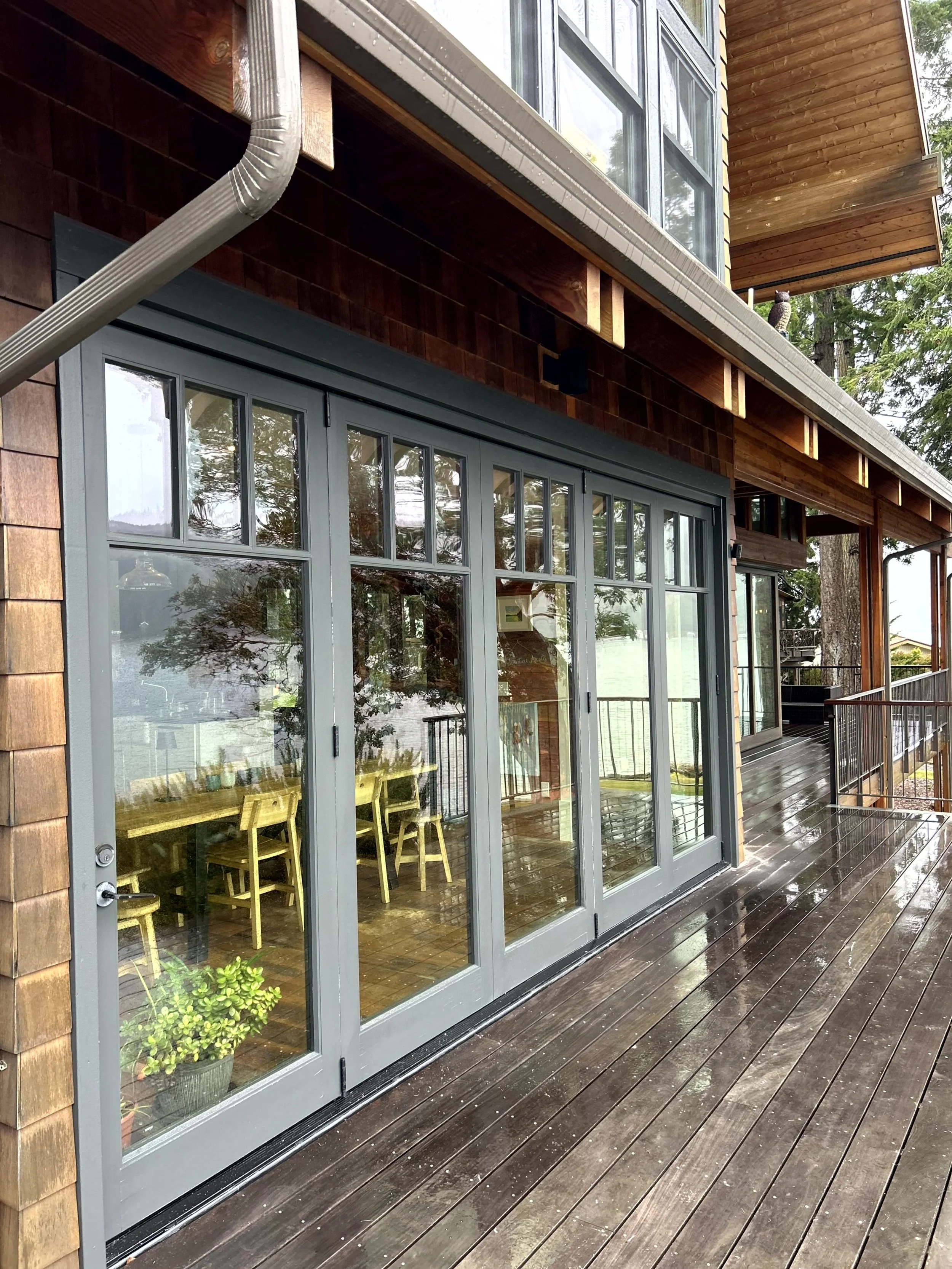 A modern house with large glass sliding doors, a wooden deck, and a brown exterior with wood shingles. There are reflections of trees in the glass.