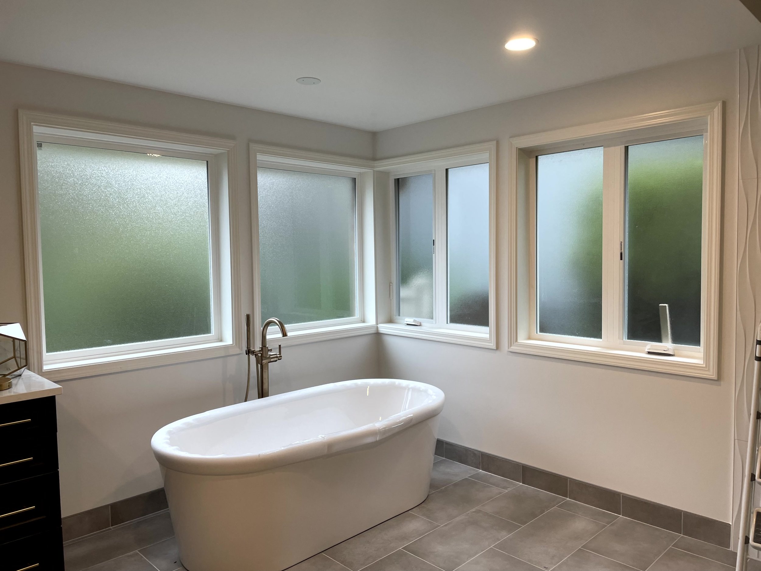 Modern bathroom with large frosted windows, a white freestanding bathtub, gray tile floor, and a small black cabinet.
