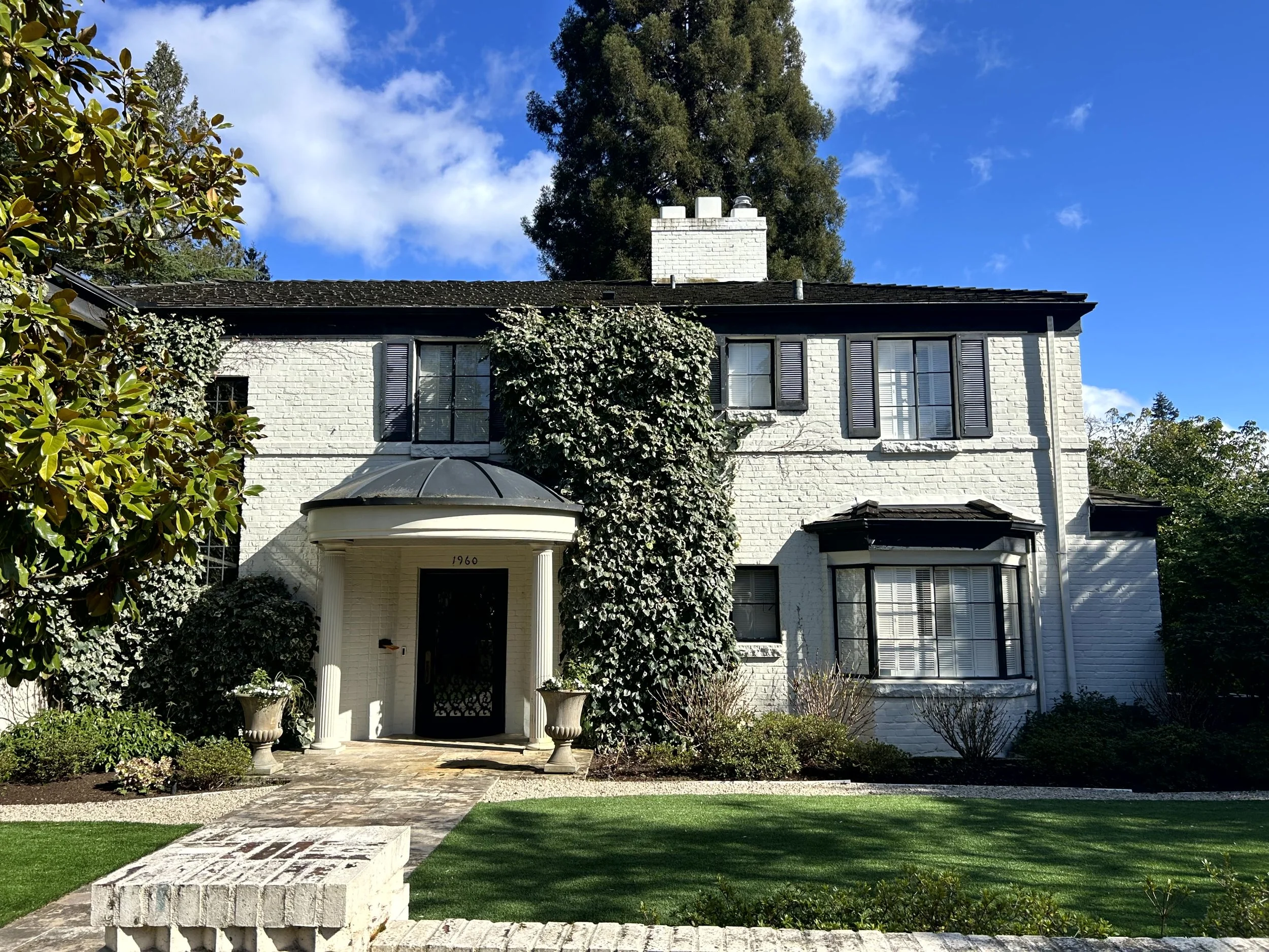 A white two-story house with black shutters, a rounded porch entrance, and a brick pathway leading to the door. The house has a garden with trimmed bushes and shrubs, and large trees behind it under a partly cloudy blue sky.