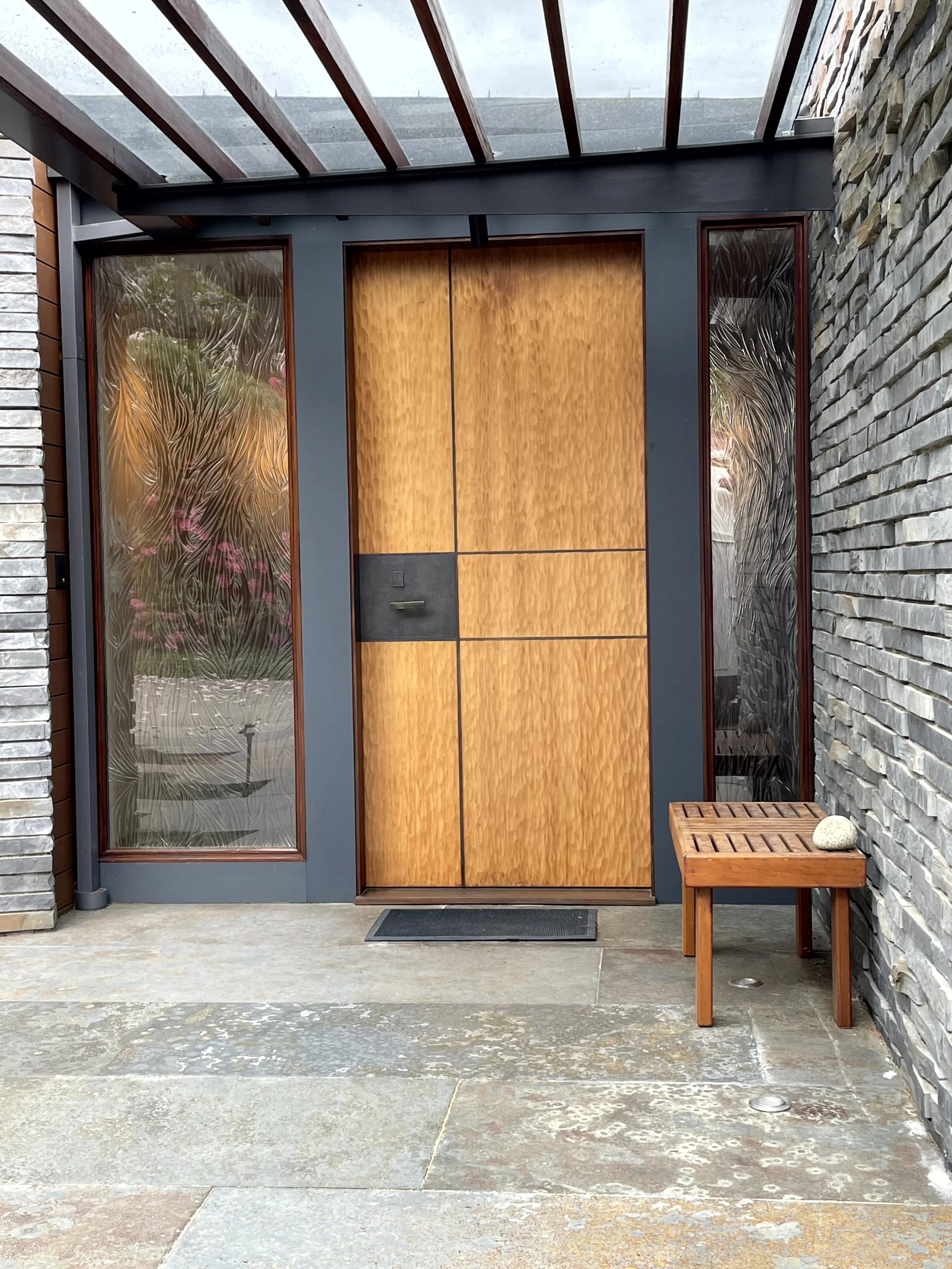 Modern front entrance with a wooden door, glass side panels with textured patterns, a small wooden bench with a spherical stone on it, and stone walls on either side.