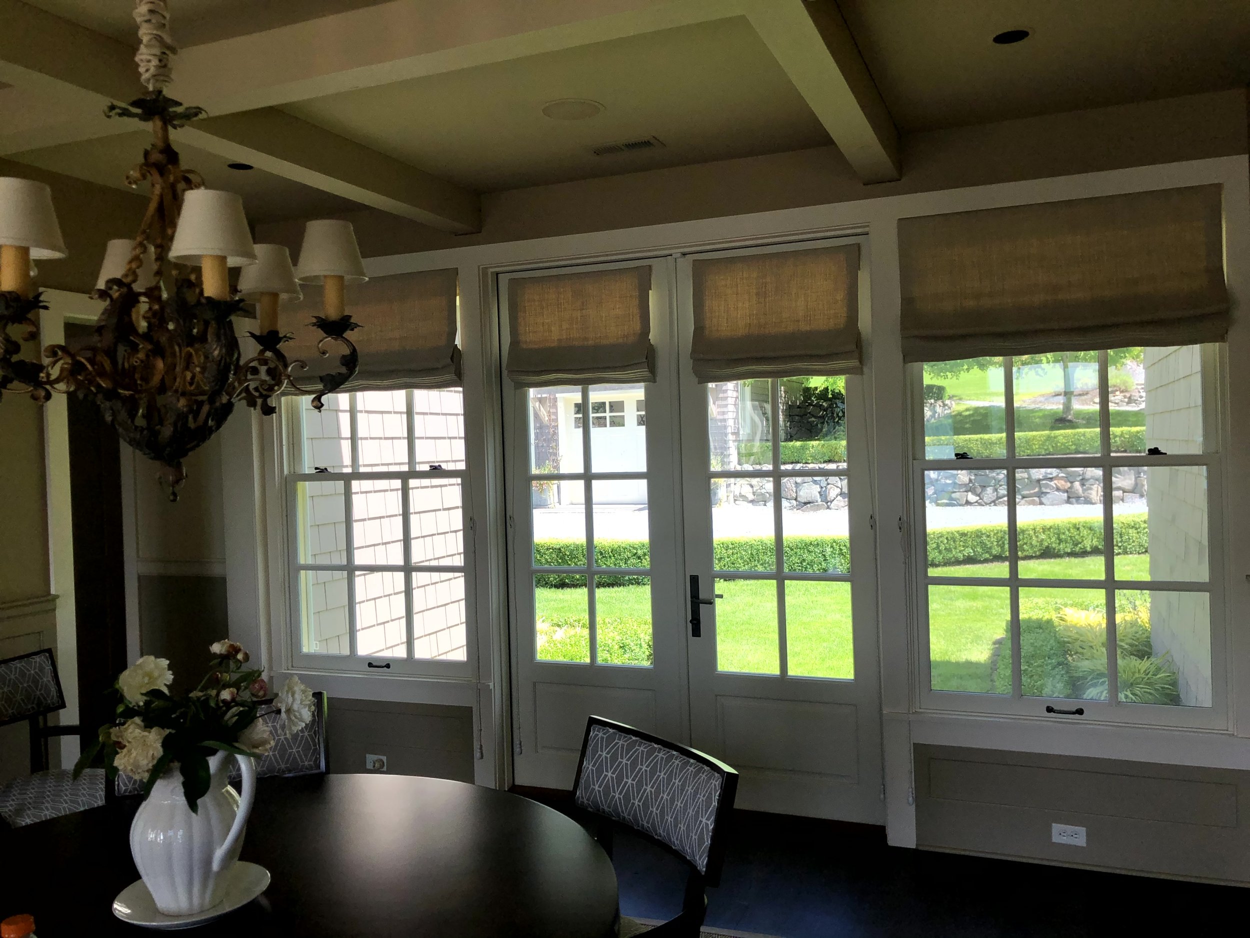 Interior view of a dining room with large windows showing a green backyard, a chandelier hanging from the ceiling, and a table with a vase of flowers.