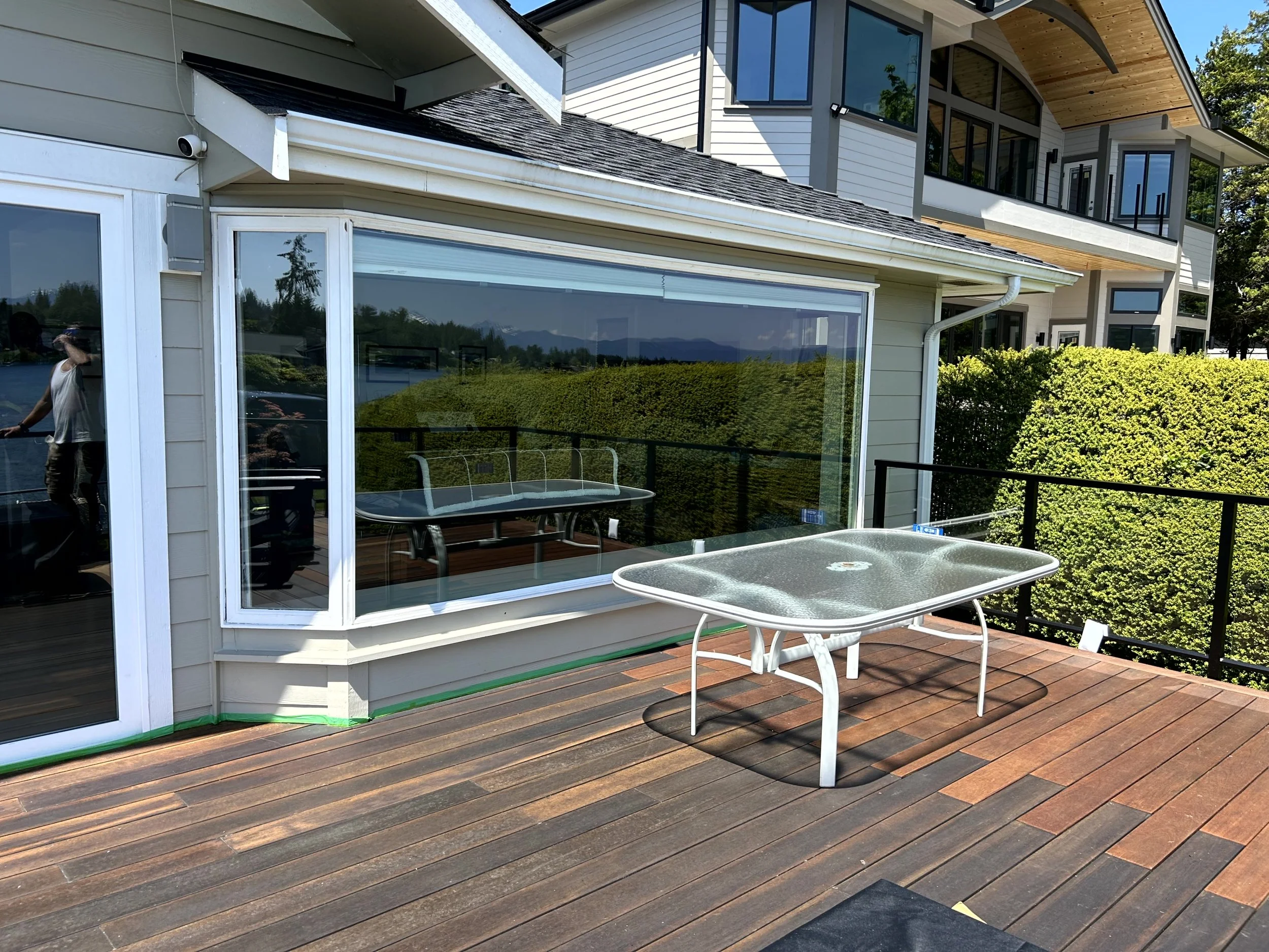 A modern house with a wooden deck outdoor patio featuring a glass-top table. Large windows reflect the lawn and sky. A person is seen in the reflection on the sliding glass door. The house has cream siding, black railings, and multiple levels with ba