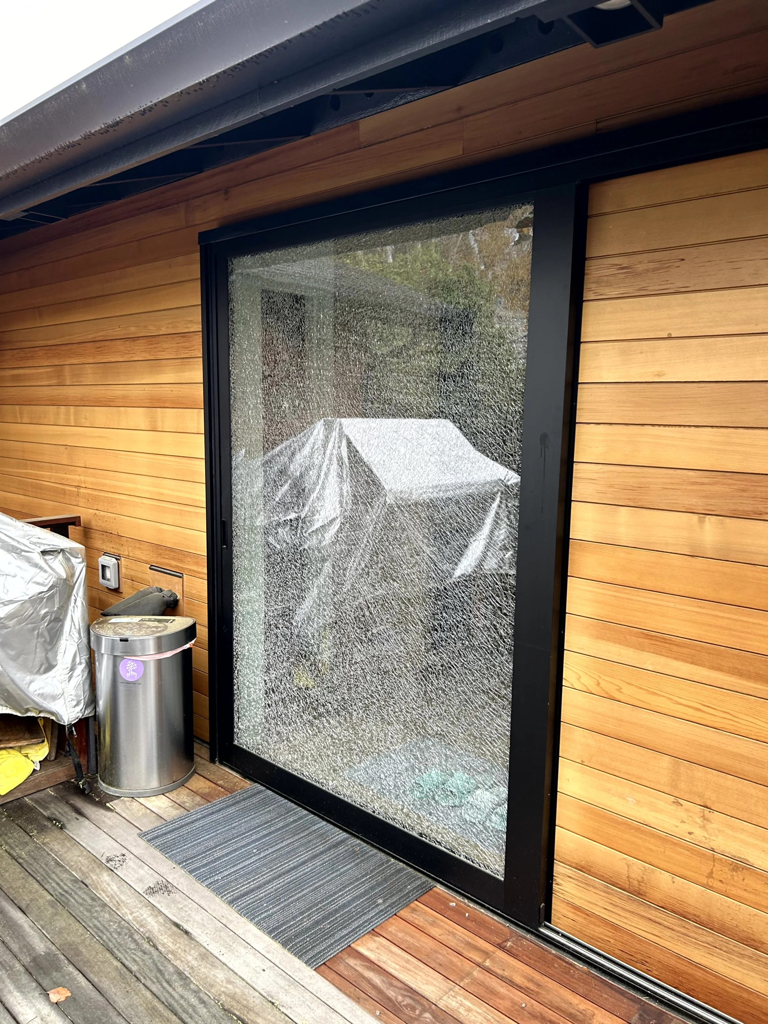 A sliding glass door with a shattered glass pane, showing cracks and broken glass, on a wooden deck outside a house with wood siding.