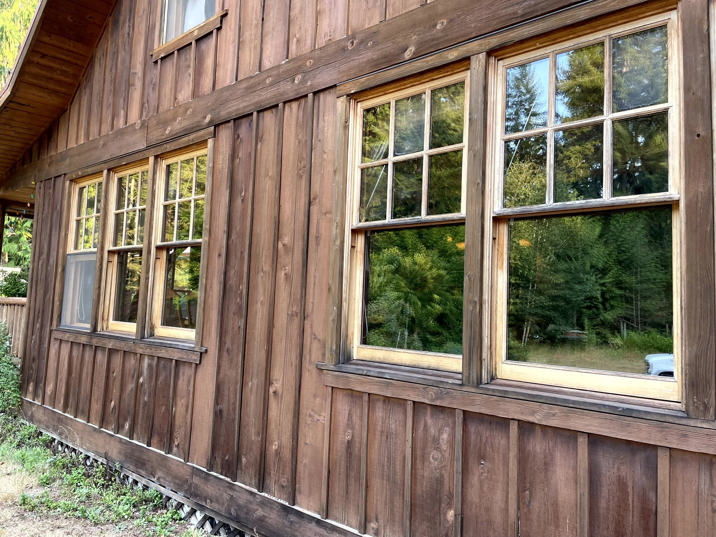 Close-up of a rustic wooden house exterior with three large multi-pane windows reflecting greenery from the outdoors.