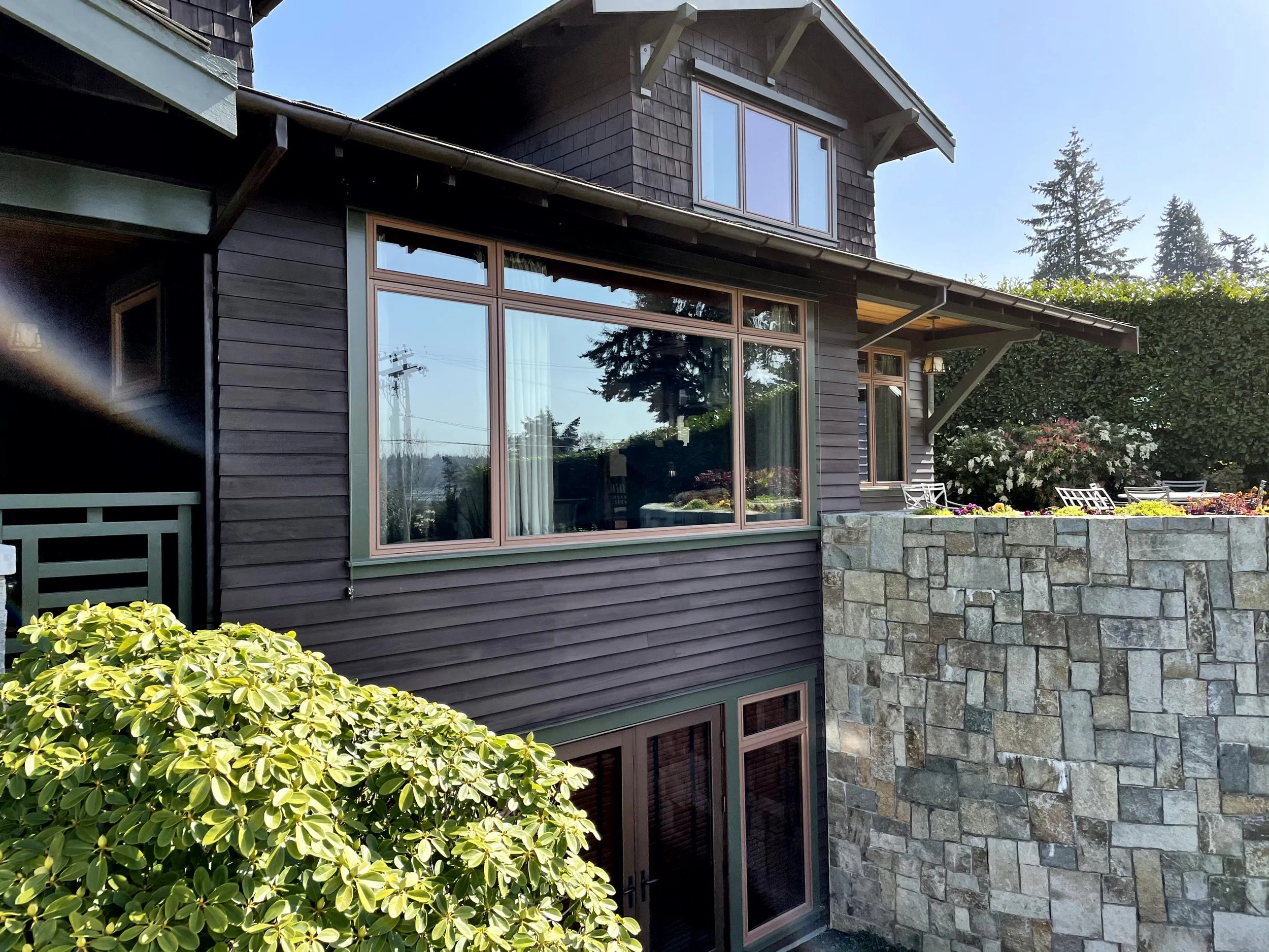 A two-story house with dark brown wood siding, large windows, and a stone retaining wall. The house is surrounded by green bushes and trees, with a bright blue sky overhead.