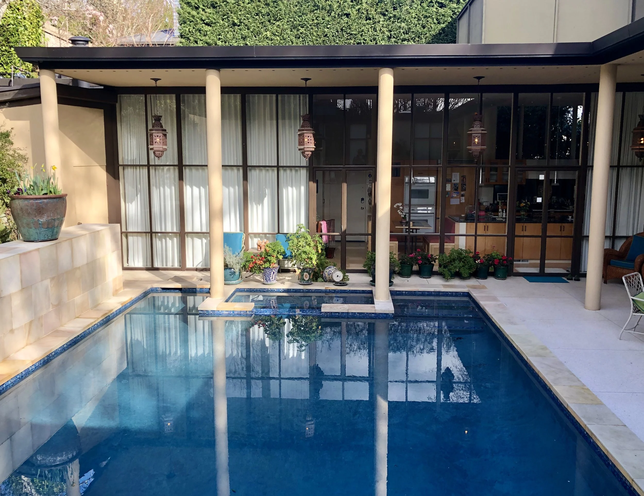 A backyard pool area with a rectangular swimming pool, potted plants along the back wall, and a house with large glass windows and sliding doors.