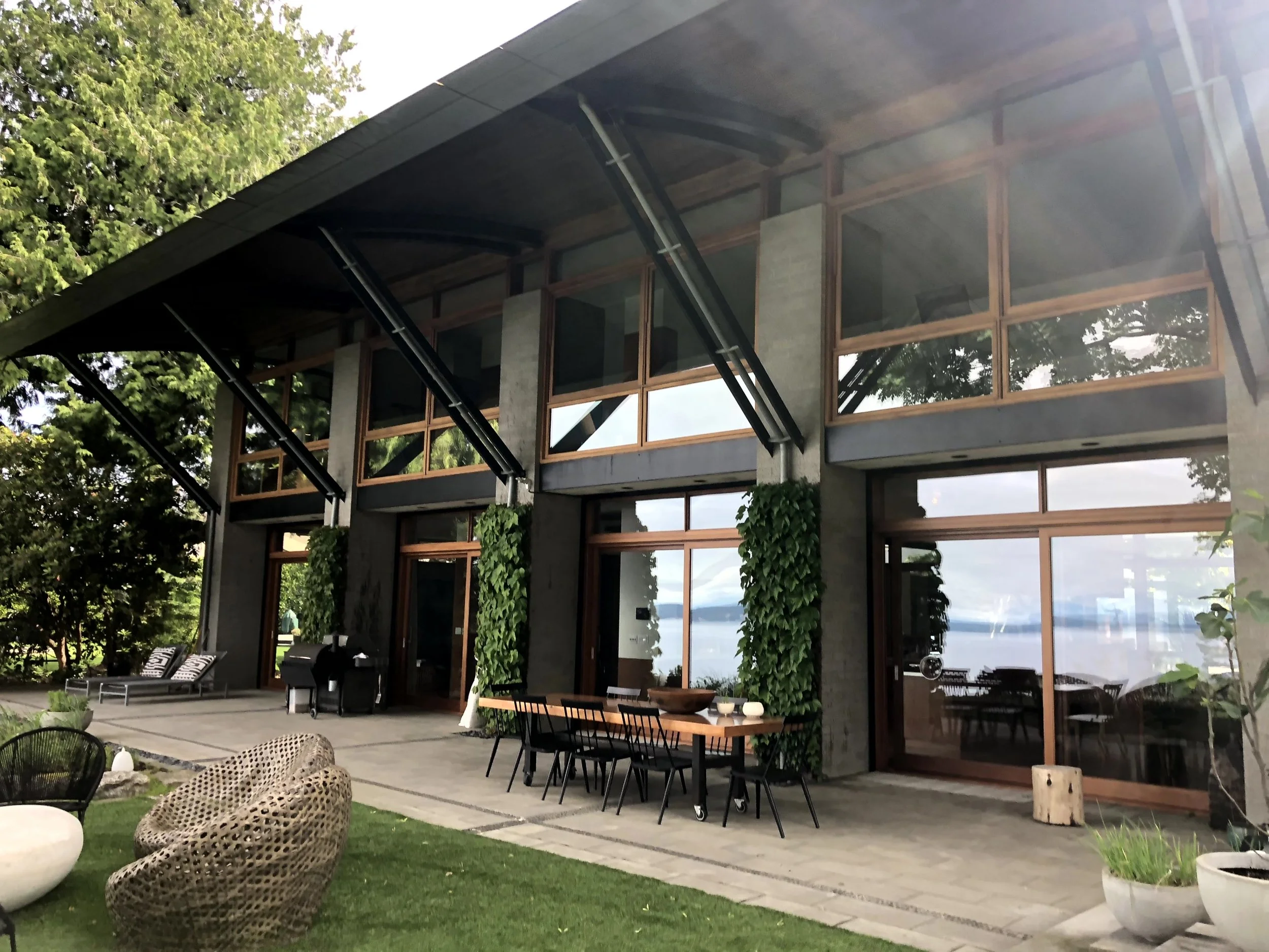 Exterior view of a modern house with large glass windows and doors, outdoor patio furniture, greenery, and a lake in the background.