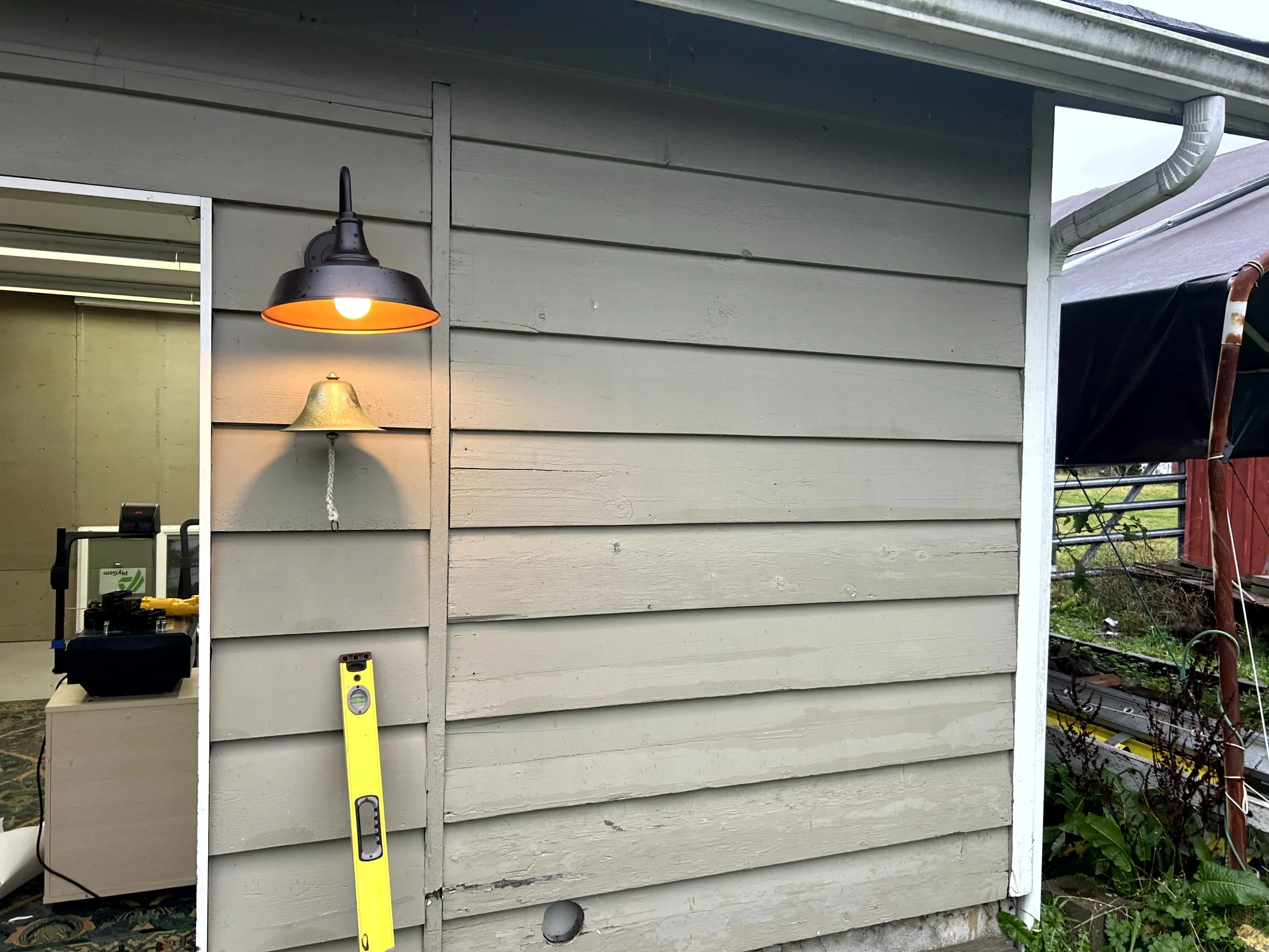 Exterior of a house with beige siding, a black and orange outdoor wall light, and a hanging bell near the light. A yellow level tool is positioned against the wall. Part of a porch or extension with visible gardening and work tools to the right.