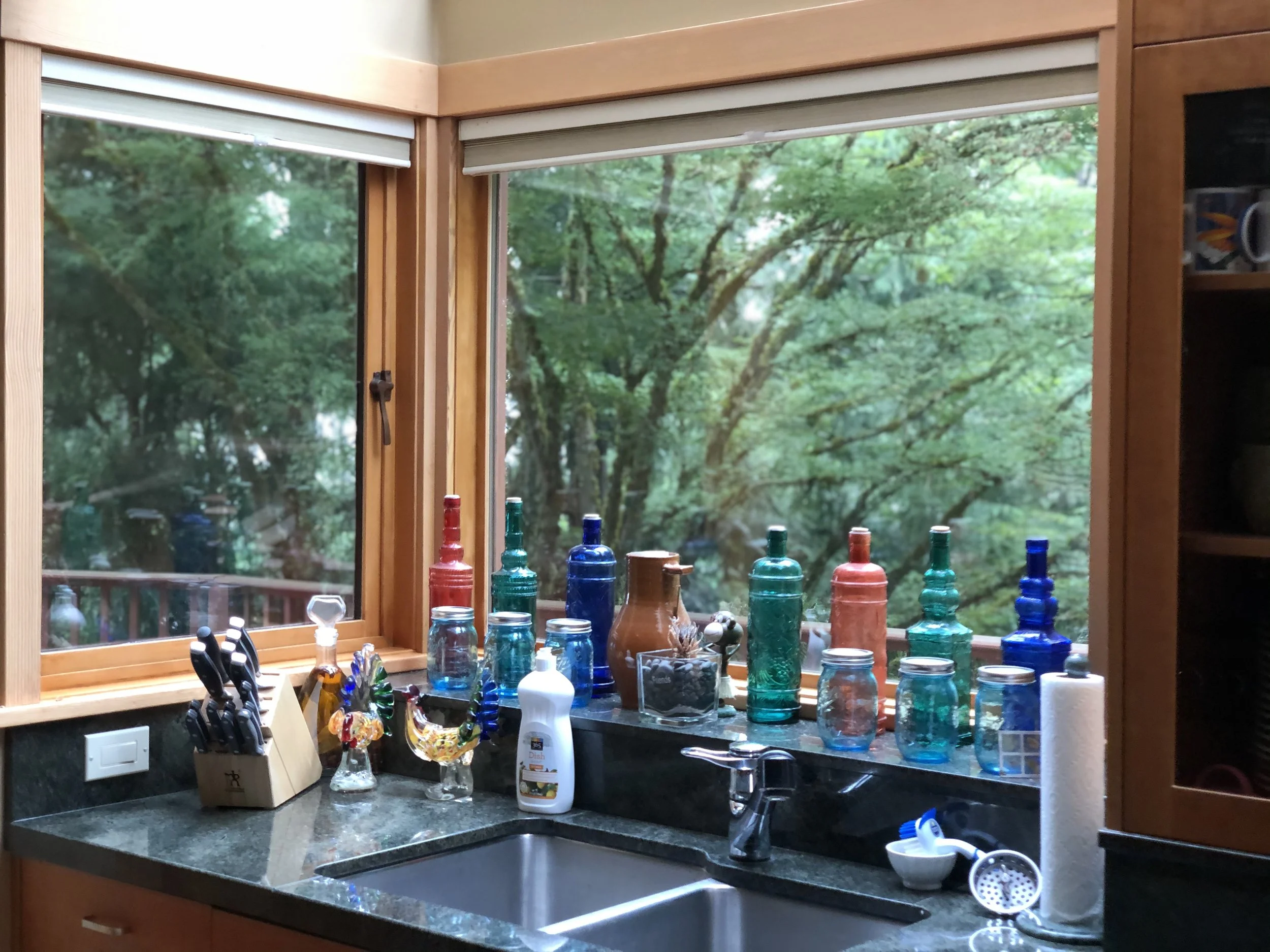 Kitchen window scene with numerous colorful glass bottles on the windowsill, a sink, knives in a knife block, and a paper towel roll, overlooking a lush green outdoor area.