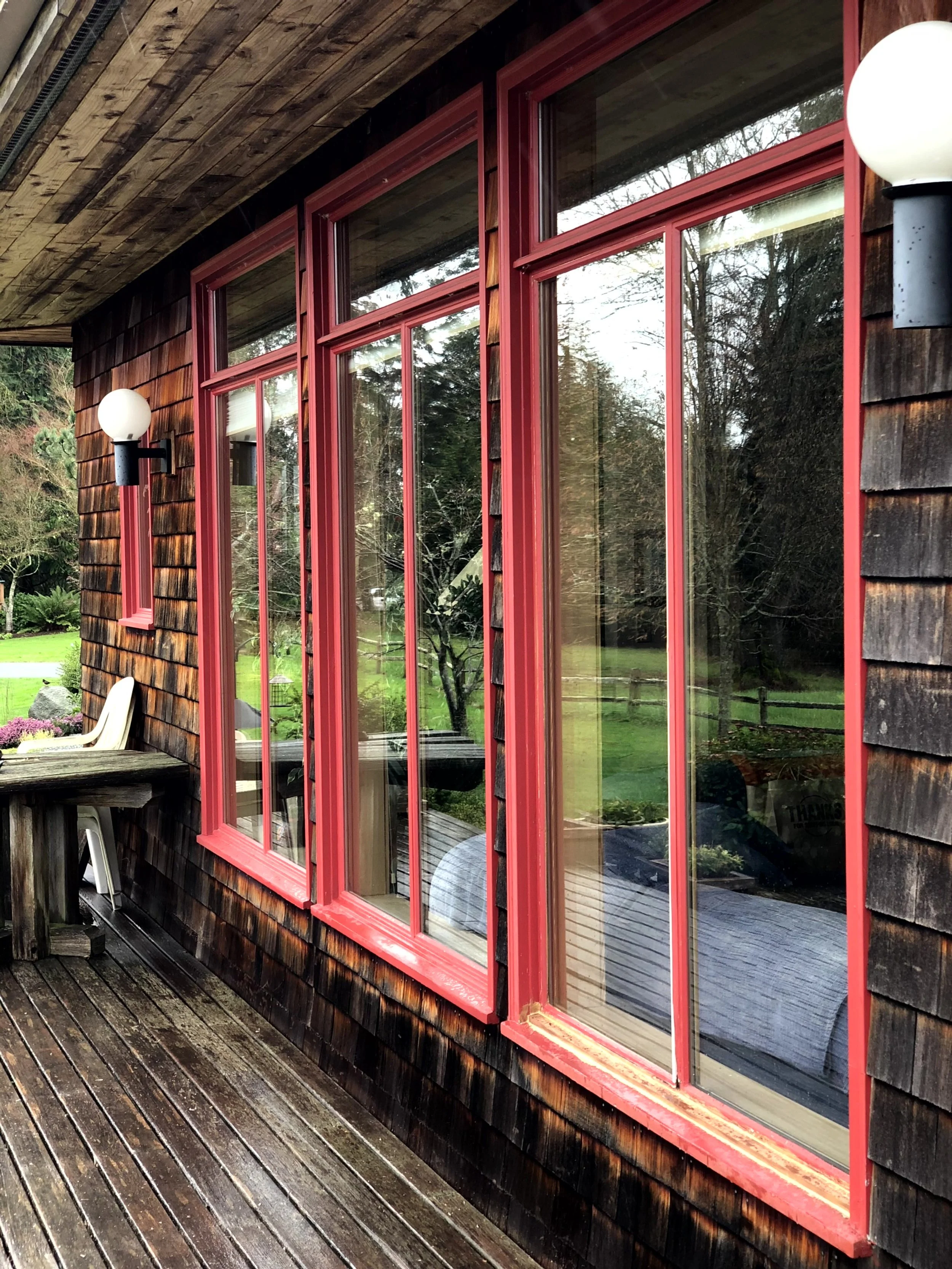 Close-up of a wooden house exterior with large red-framed windows and outdoor lighting fixtures, showing reflections of a garden and trees.