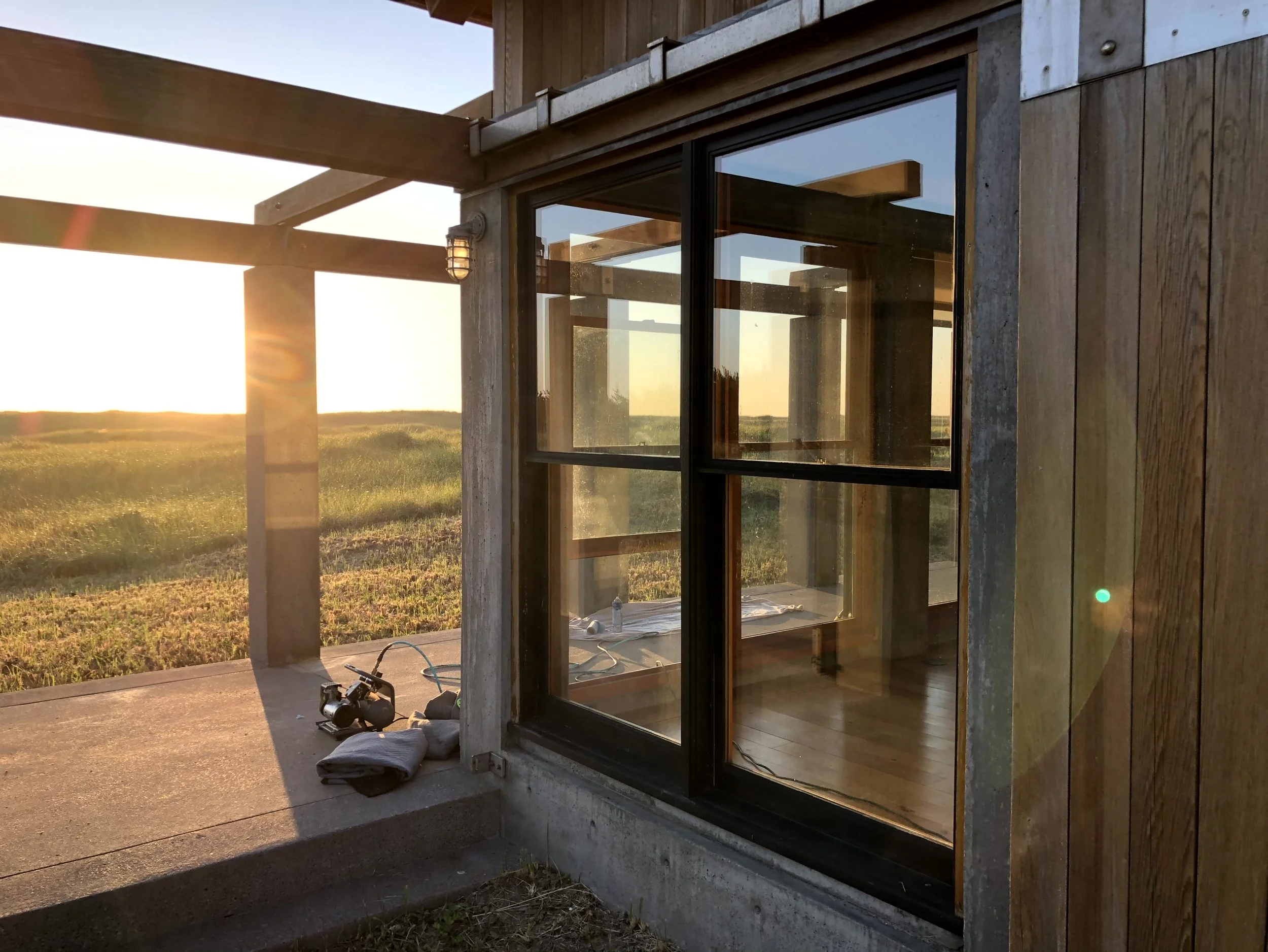 Sunset view of a modern house with large glass windows, wooden beams, and a concrete patio, overlooking an open field with green grass.