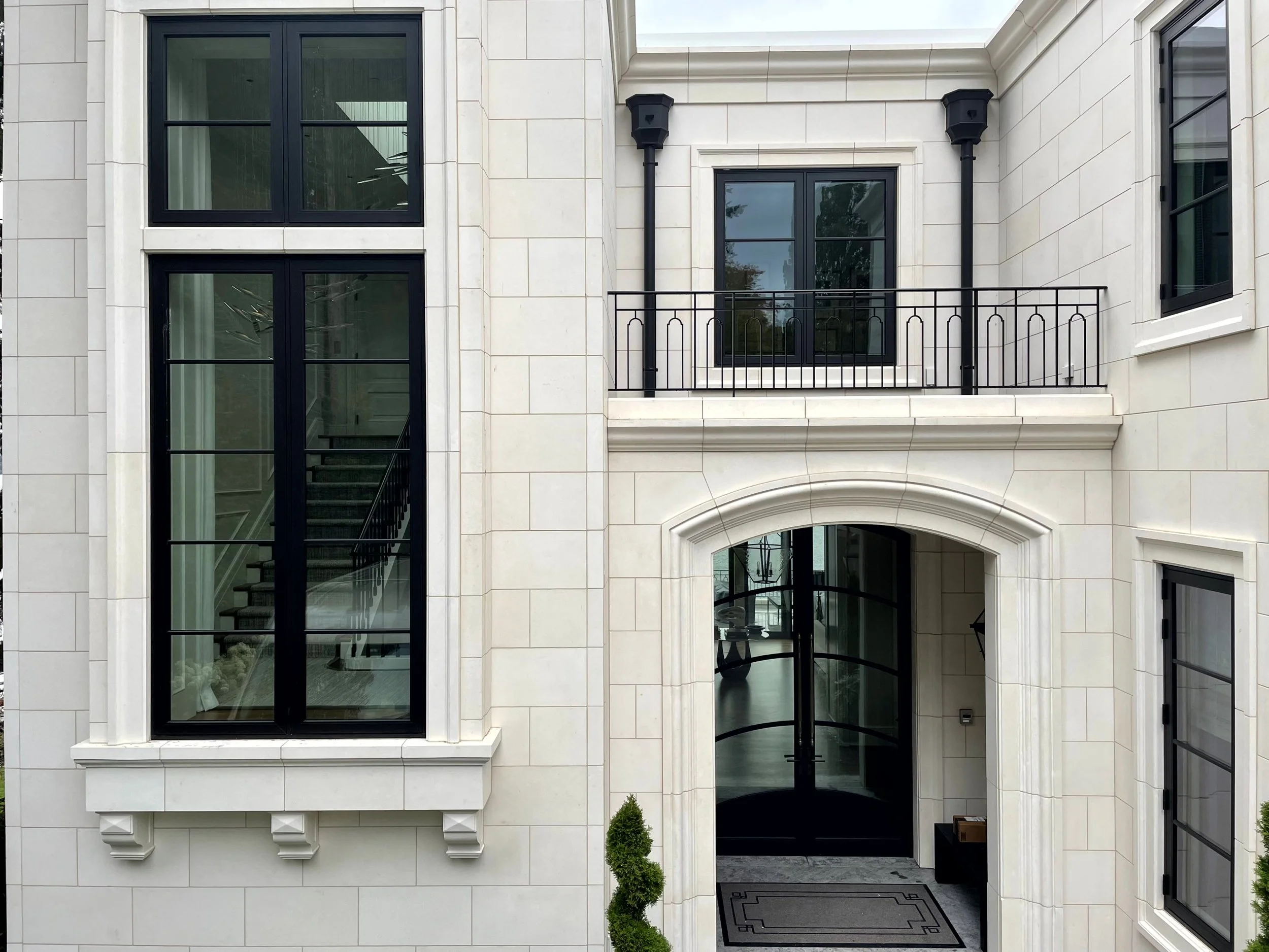 Front entrance of a modern building with large black-framed windows, a glass door, and decorative black railings on a second-floor balcony.