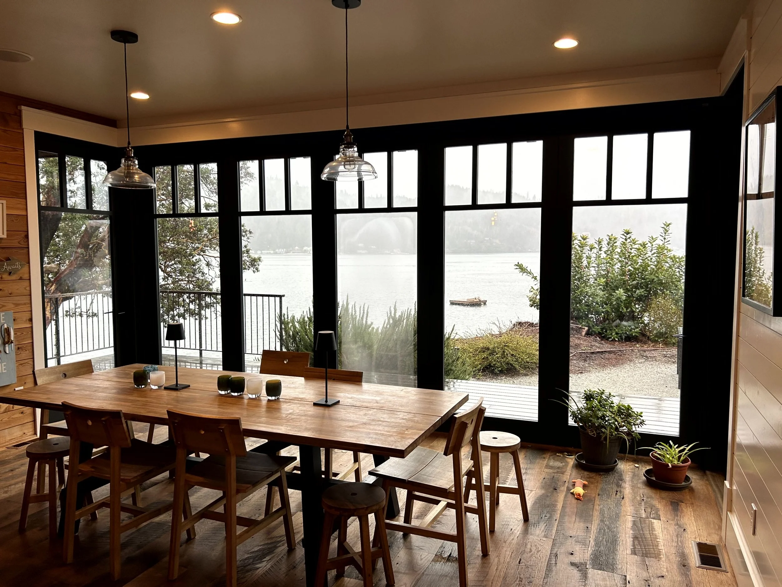 Cozy dining room with large glass windows overlooking a lake, wooden table with chairs, potted plants, and hanging pendant lights.