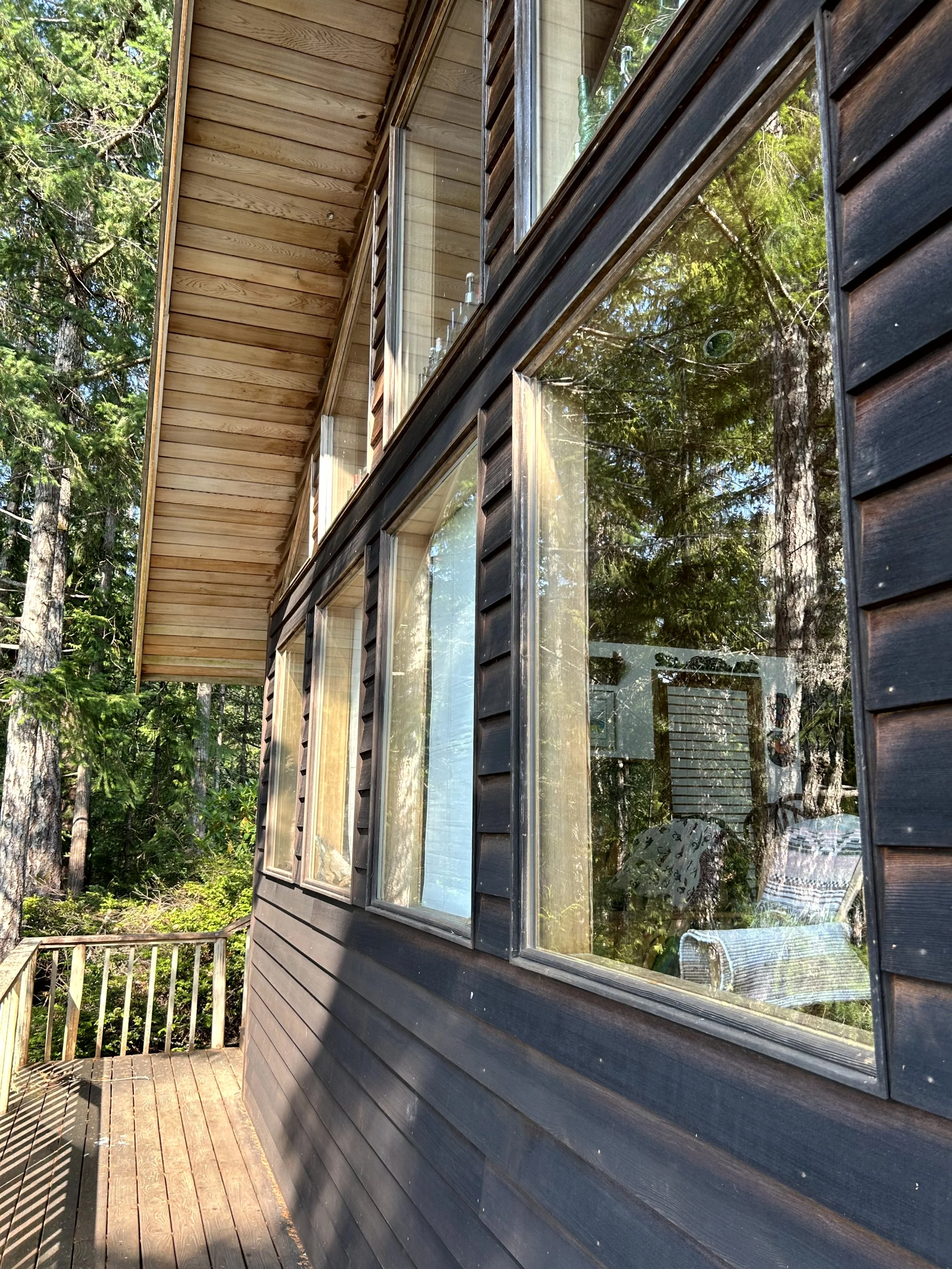 View of the side of a wooden house with large windows reflecting trees and a deck with a railing.