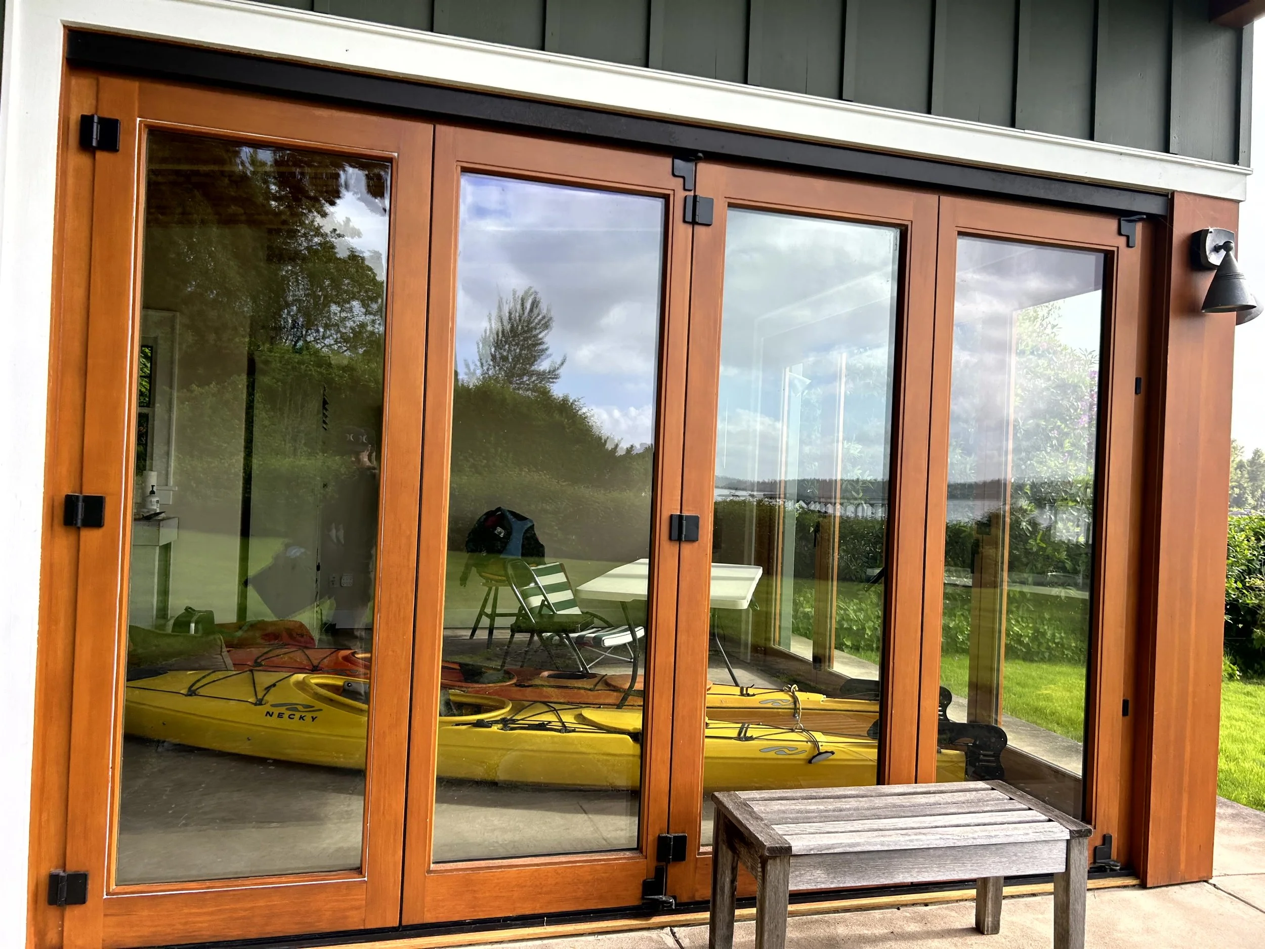 A glass patio door with wooden frame, showing a yellow kayak inside, outside a backyard with green grass, bushes, trees, and a cloudy sky.