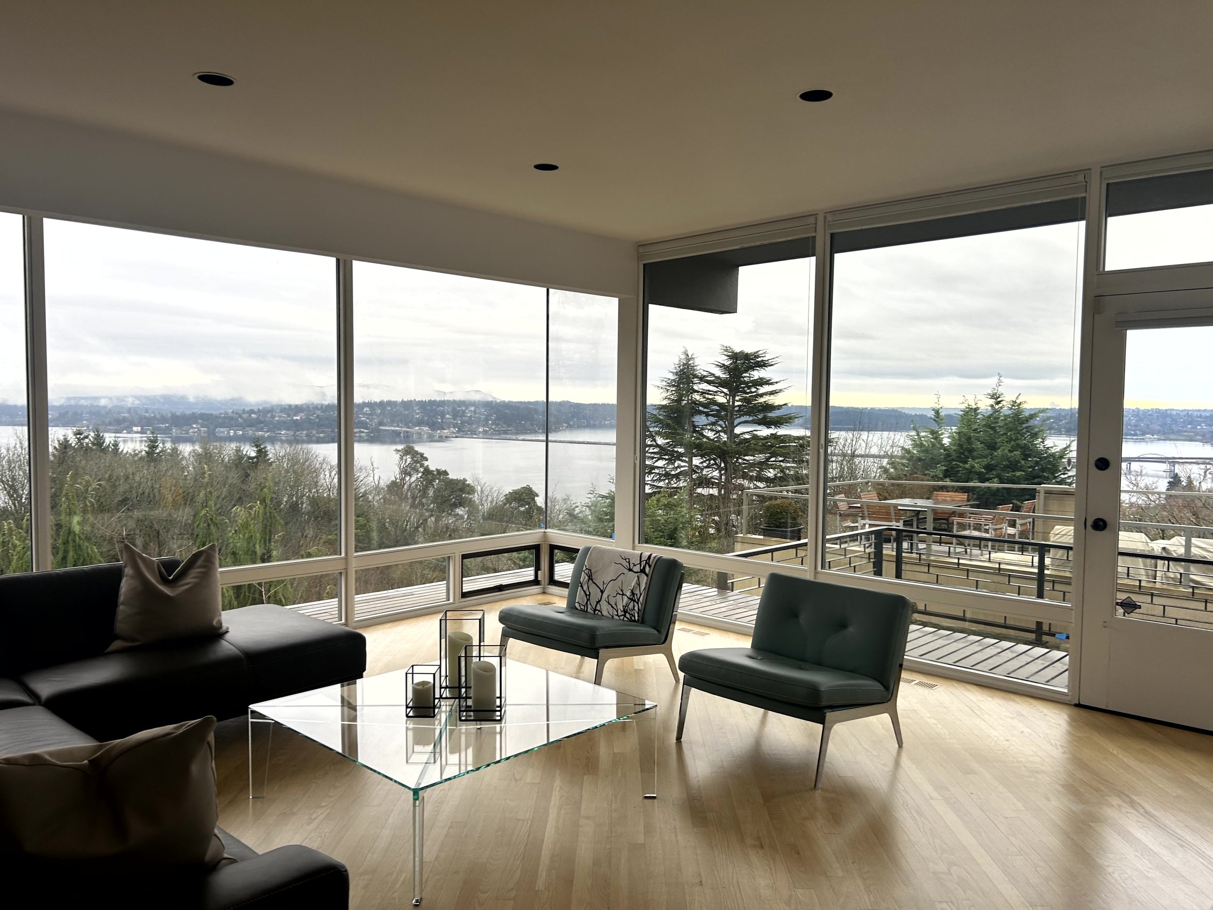 Modern living room with large floor-to-ceiling windows overlooking a lake and trees, furnished with a black leather sofa, two teal chairs, and a glass coffee table with candles, with a wood floor and sliding door leading to a balcony.