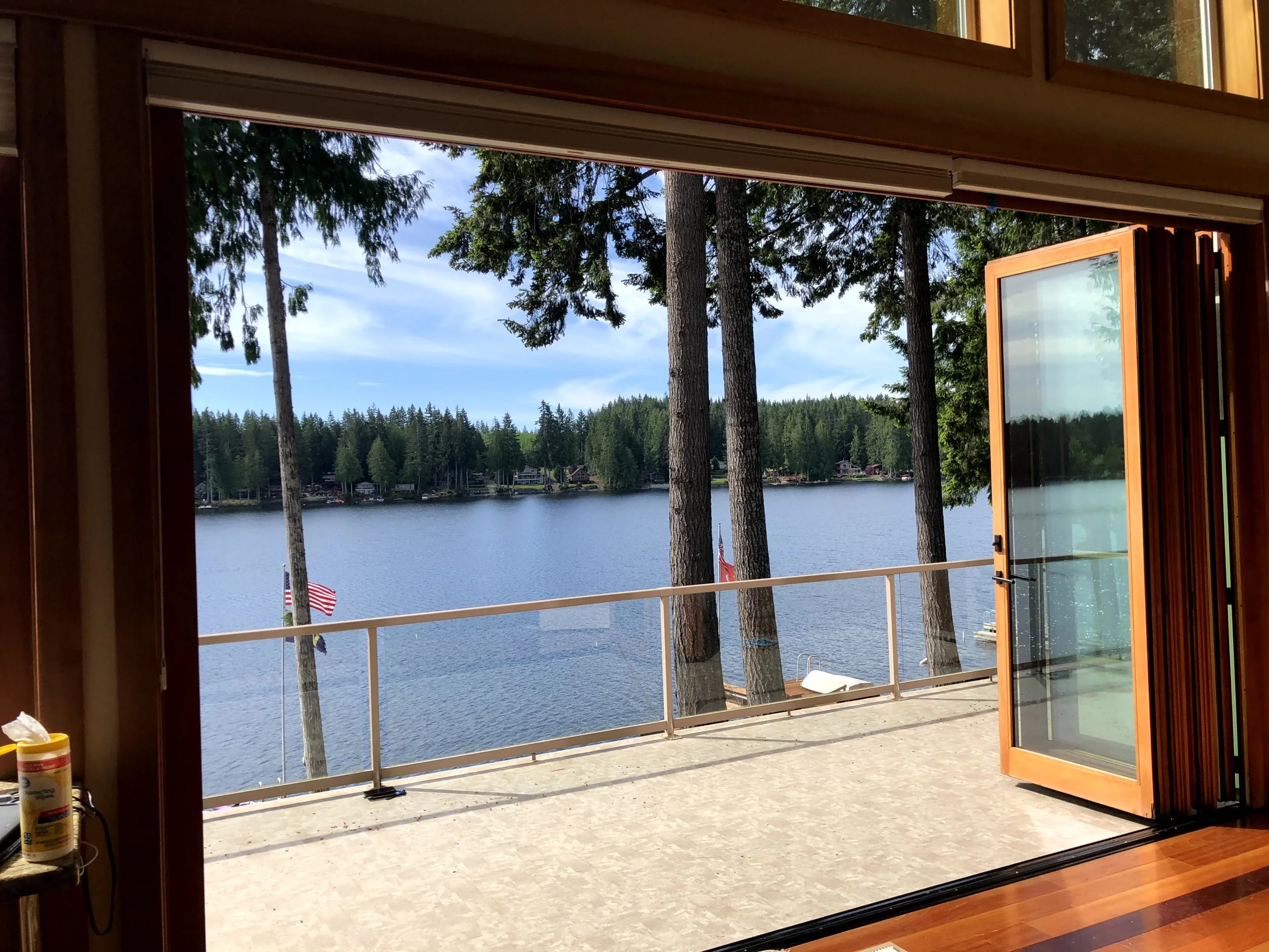 View of a lake through open glass doors from a room with a wooden floor. The balcony has a safety railing, and there are trees, water, and houses across the lake. The sky is partly cloudy.