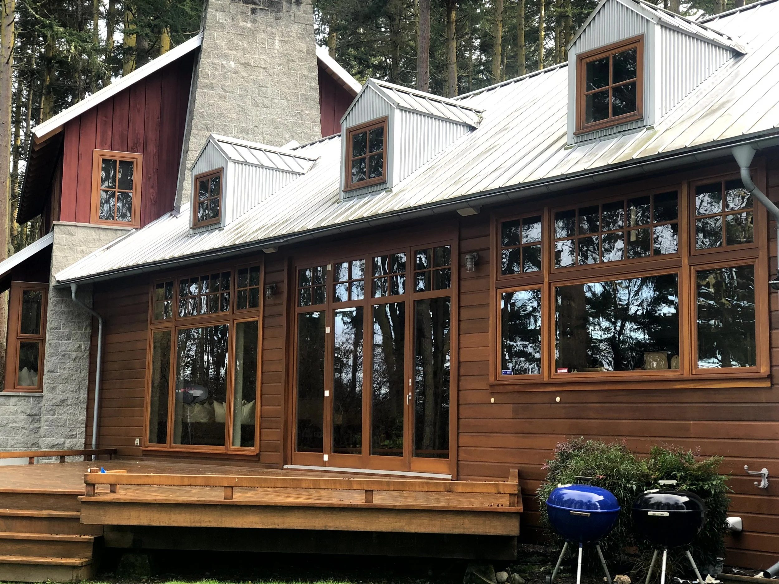 A house with large windows, a wooden deck, and a metal roof, surrounded by trees.