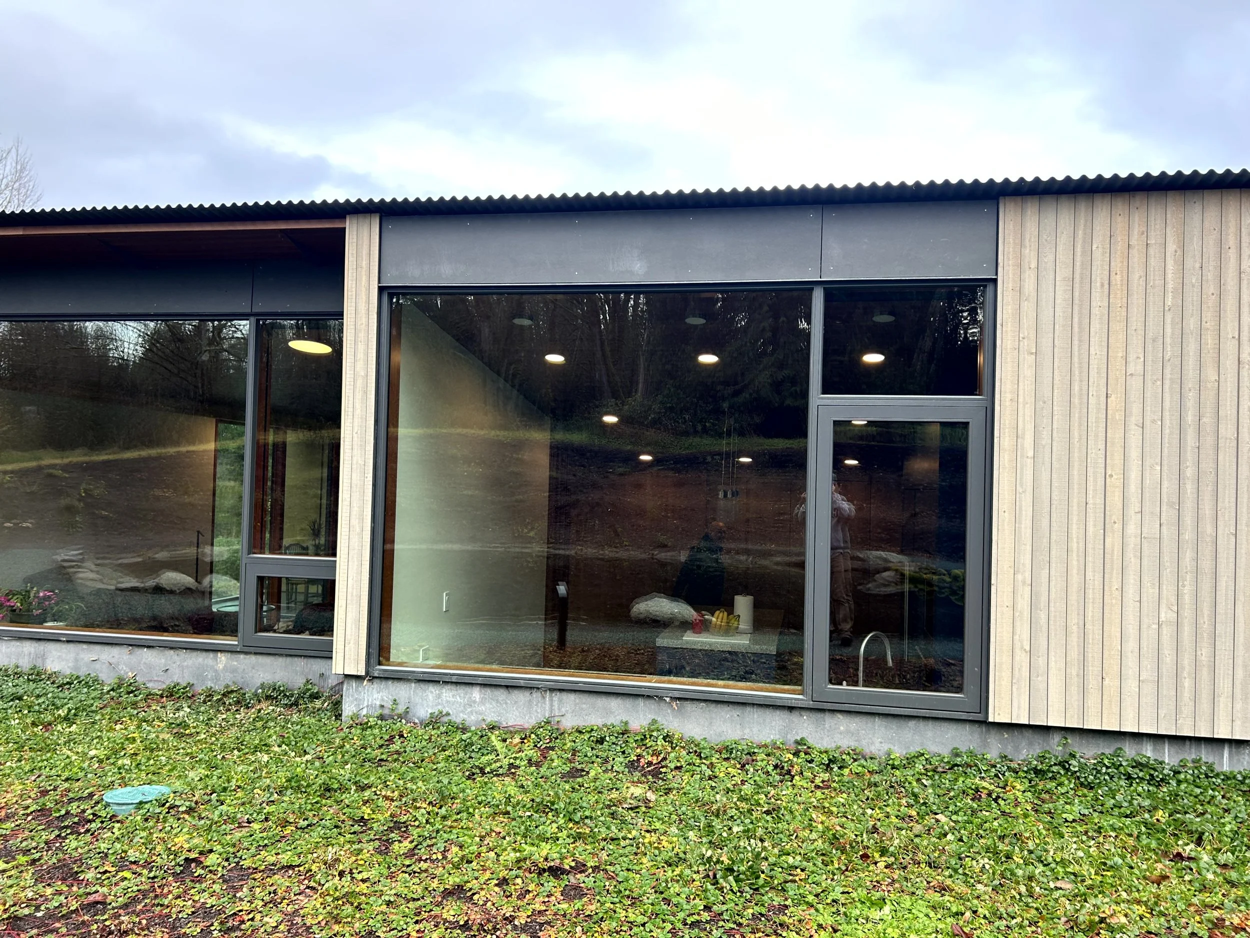 Exterior of a modern building with large glass windows and wooden siding, surrounded by green ground cover plants.