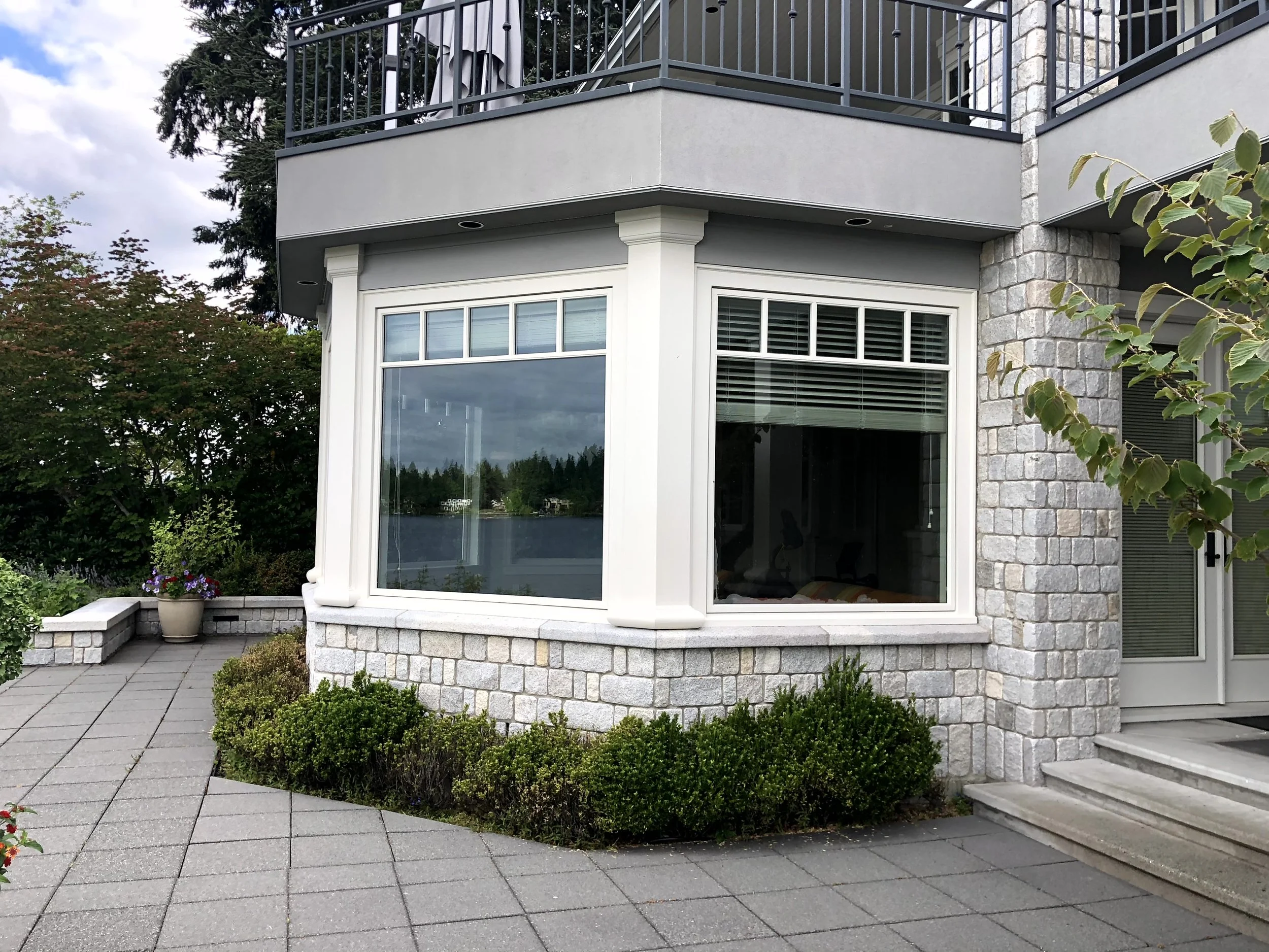 Exterior view of a house with large front windows, stone and gray stucco walls, a small front porch with steps, and a landscaped garden with shrubs and potted flowers.