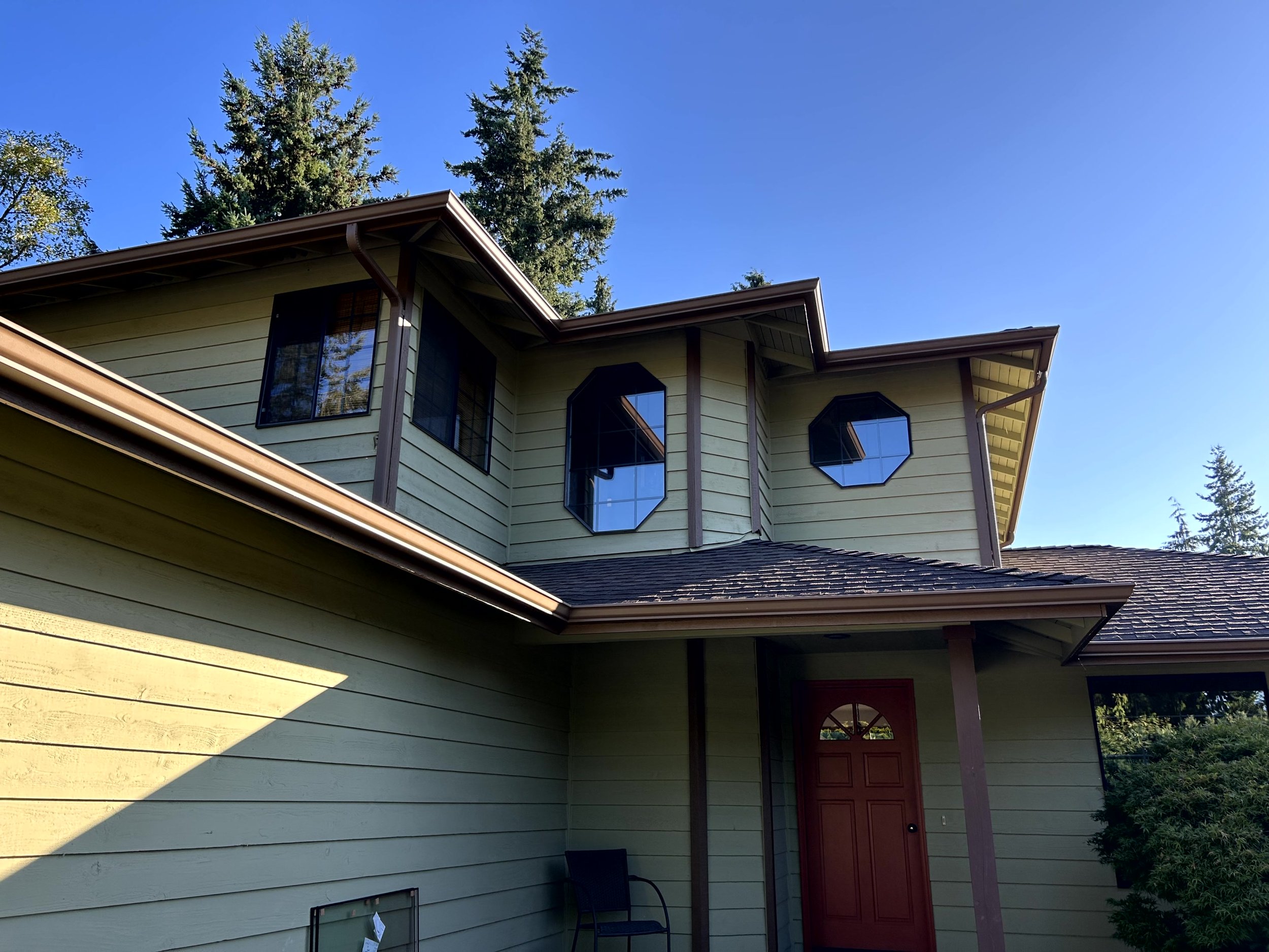 A two-story house with light yellow horizontal siding, a brown door, and octagonal and rectangular windows, surrounded by tall evergreen trees under a clear blue sky.